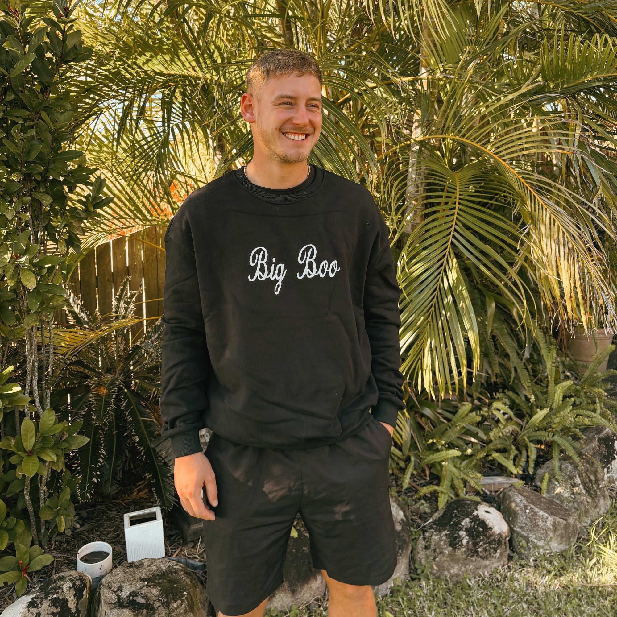 Man in a black Big Boo embroidered crew sweatshirt and black shorts standing in a tropical garden