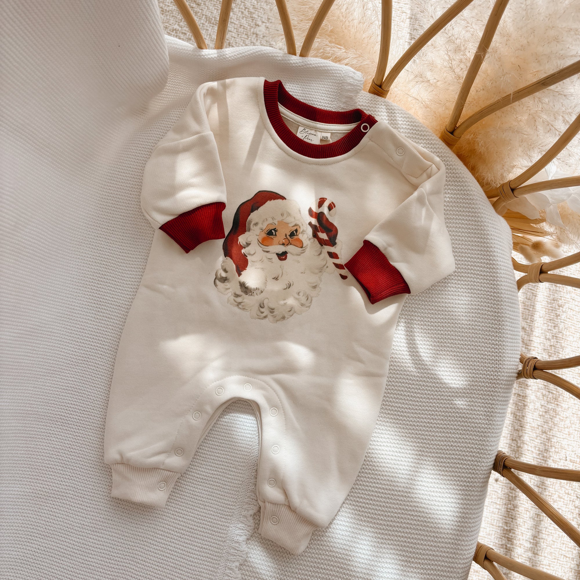 Cream baby jumpsuit with a vintage Santa print and red contrast cuffs, flat lay in a bassinet