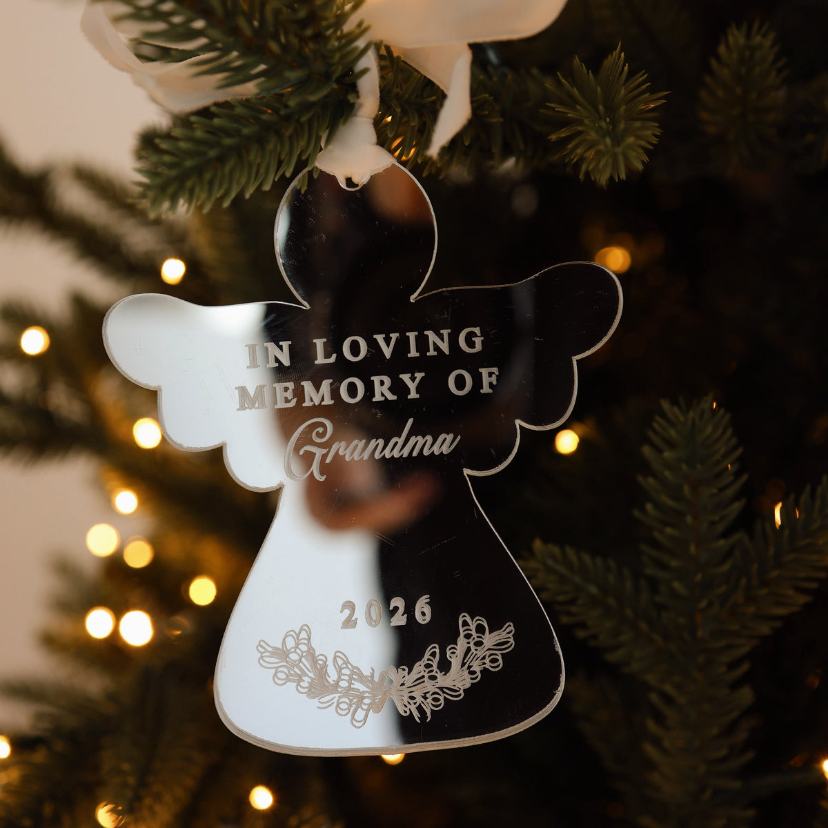 Mirrored acrylic angel ornament engraved 'In Loving Memory of Grandma 2026' with a foliage motif, hung from a white ribbon on a lit tree