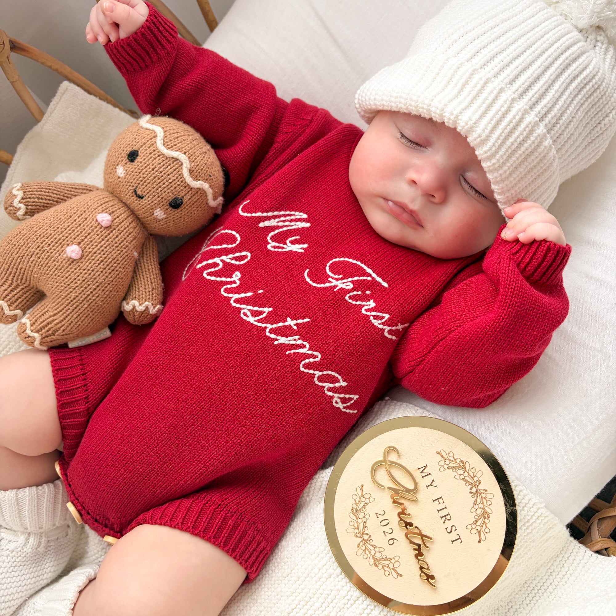 Sleeping baby in a red knit romper embroidered My First Christmas, with a gingerbread toy and gold keepsake disc