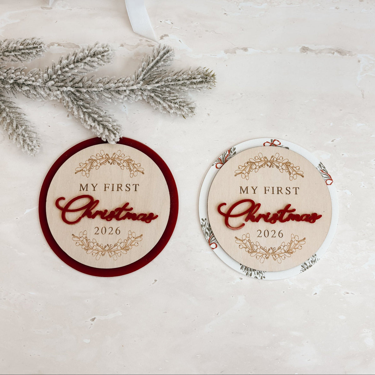 Two round Christmas gift tags with 'MY FIRST Christmas 2026' text, one with a dark red border and one with a white border, displayed on a light textured surface with a kraft gift box and silver pine branches.