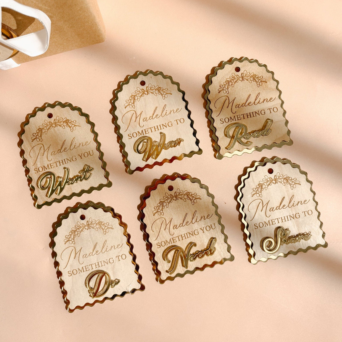 Six decorative gift tags with scalloped edges and gold script text reading 'Madeline Something to [adjective]' arranged on a warm beige surface beside wrapped brown paper gift boxes with white ribbons.