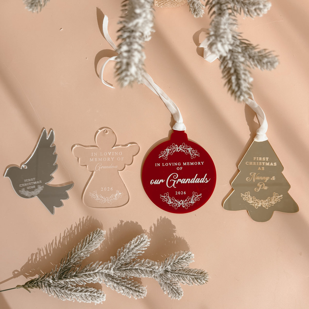 Flat lay of four personalised acrylic Christmas ornaments: mirrored dove, frosted angel and red circle memorial keepsakes, and a gold tree reading 'First Christmas as Nanny &amp; Pa'