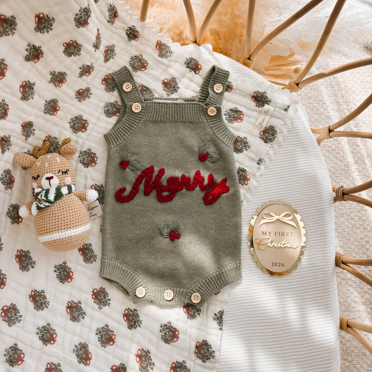 Sage green Merry knit overall romper flat lay with a crochet reindeer rattle and a gold My First Christmas 2026 keepsake disc