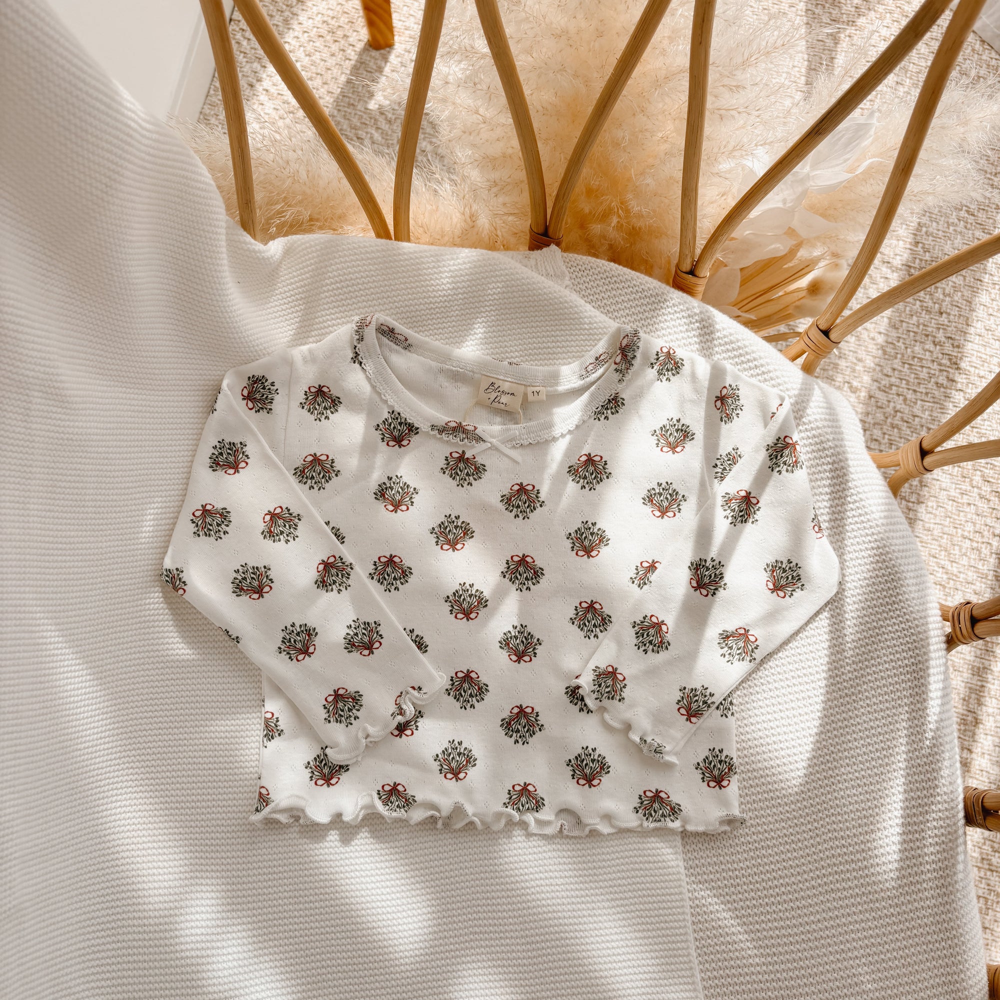 Cream long sleeve baby top with a wreath print and lettuce edge hem, flat lay in a bassinet