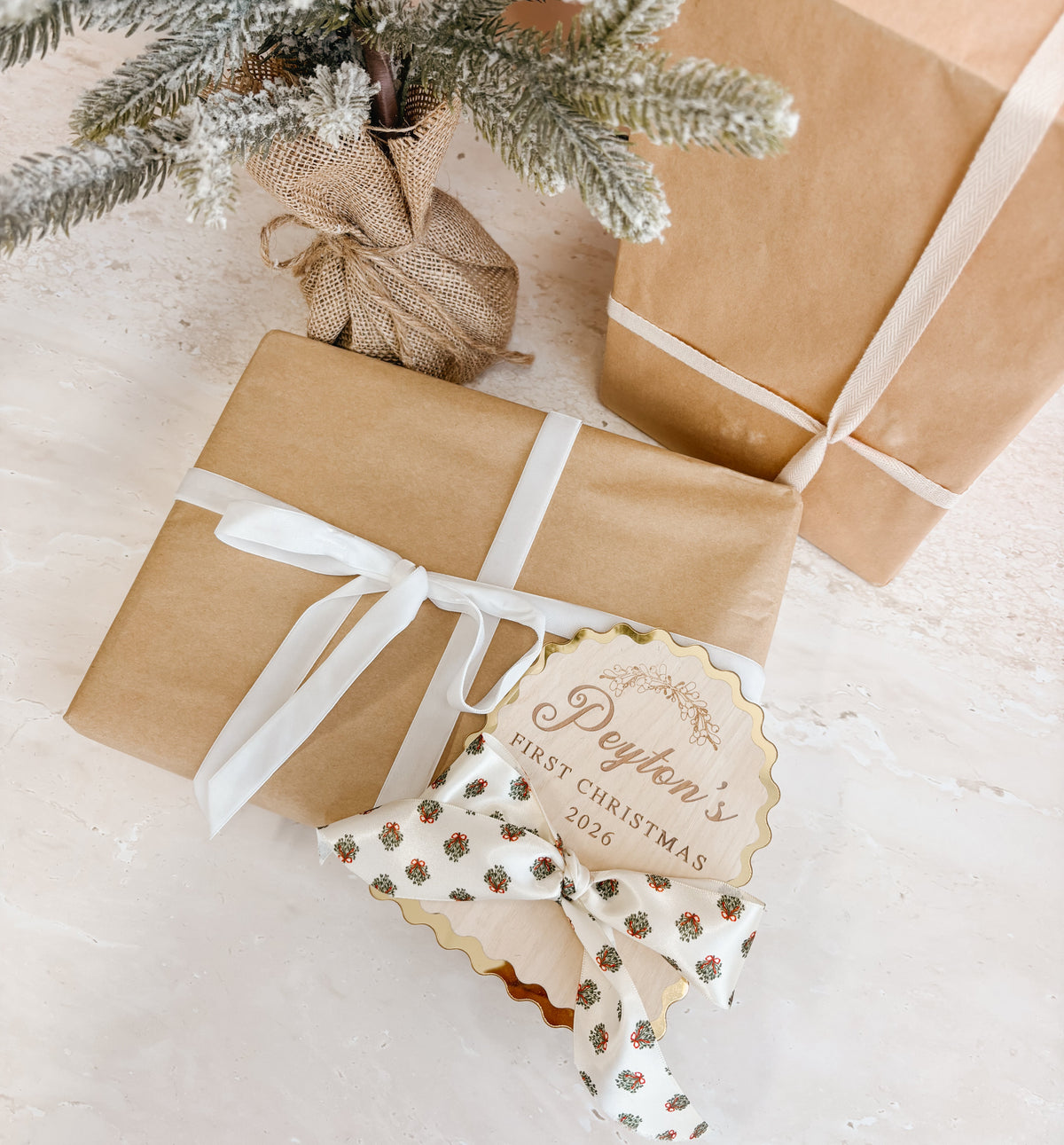 A styled flat-lay of wrapped gift boxes on a light surface, with one box in the foreground tied with a white ribbon and a decorative tag reading 'Peyton's First Christmas 2026,' plus a patterned bow and frosted evergreen branches in the background.