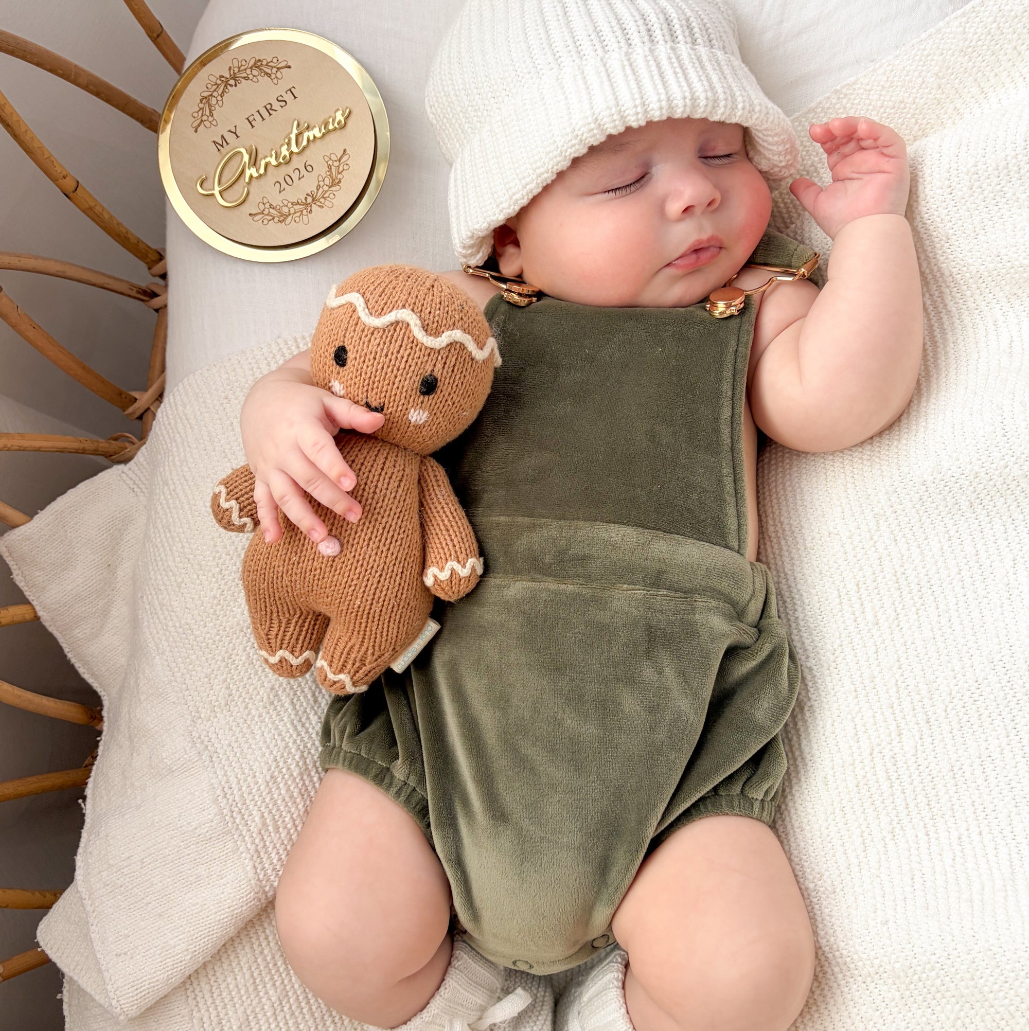 Sleeping baby in an olive velvet overall romper and white pom beanie with a knitted gingerbread toy and My First Christmas keepsake disc
