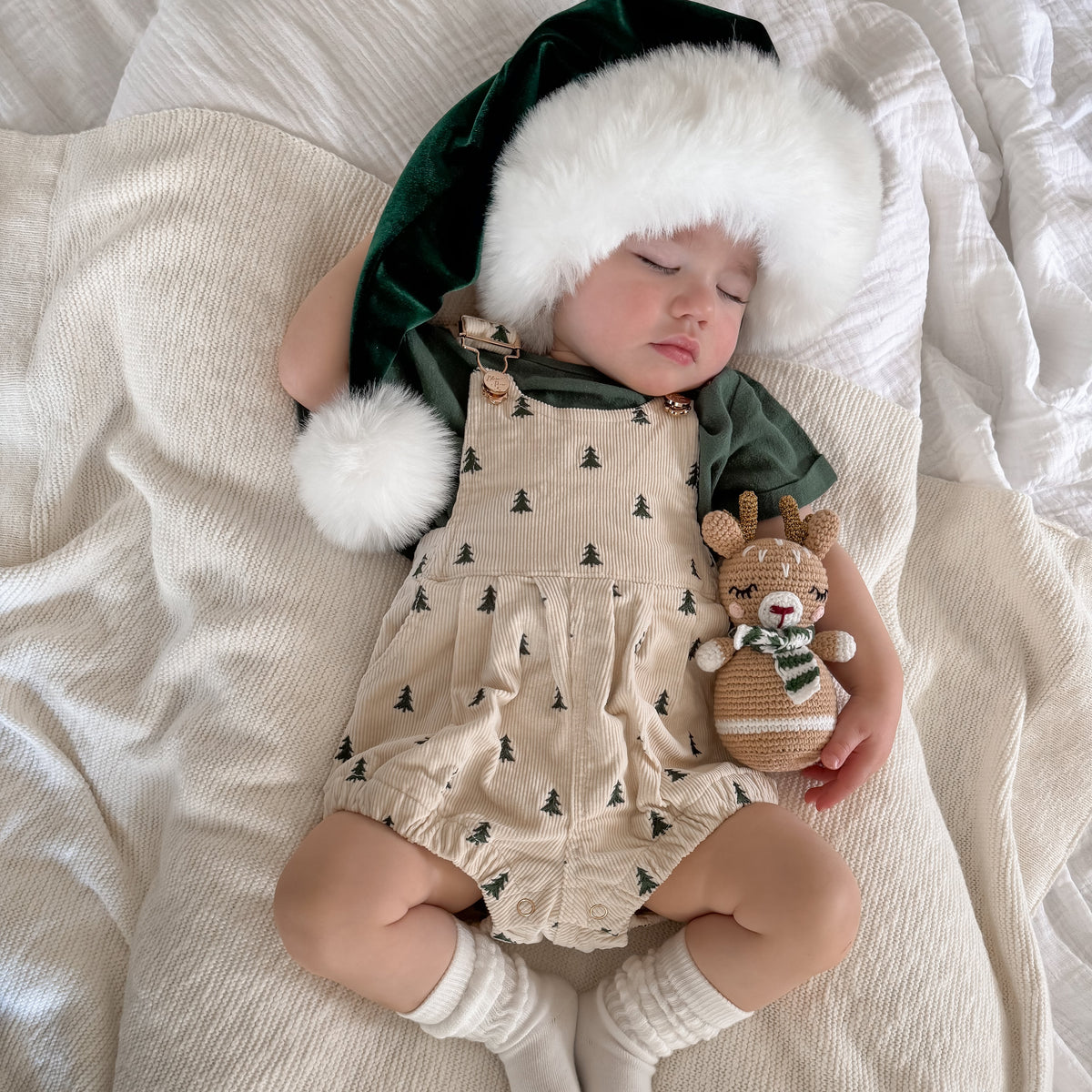 A sleeping baby wearing a festive Christmas outfit, including a dark green velvet Santa hat with white faux fur trim, beige patterned overalls, and white socks, lying on a textured cream blanket and holding a knitted reindeer toy.