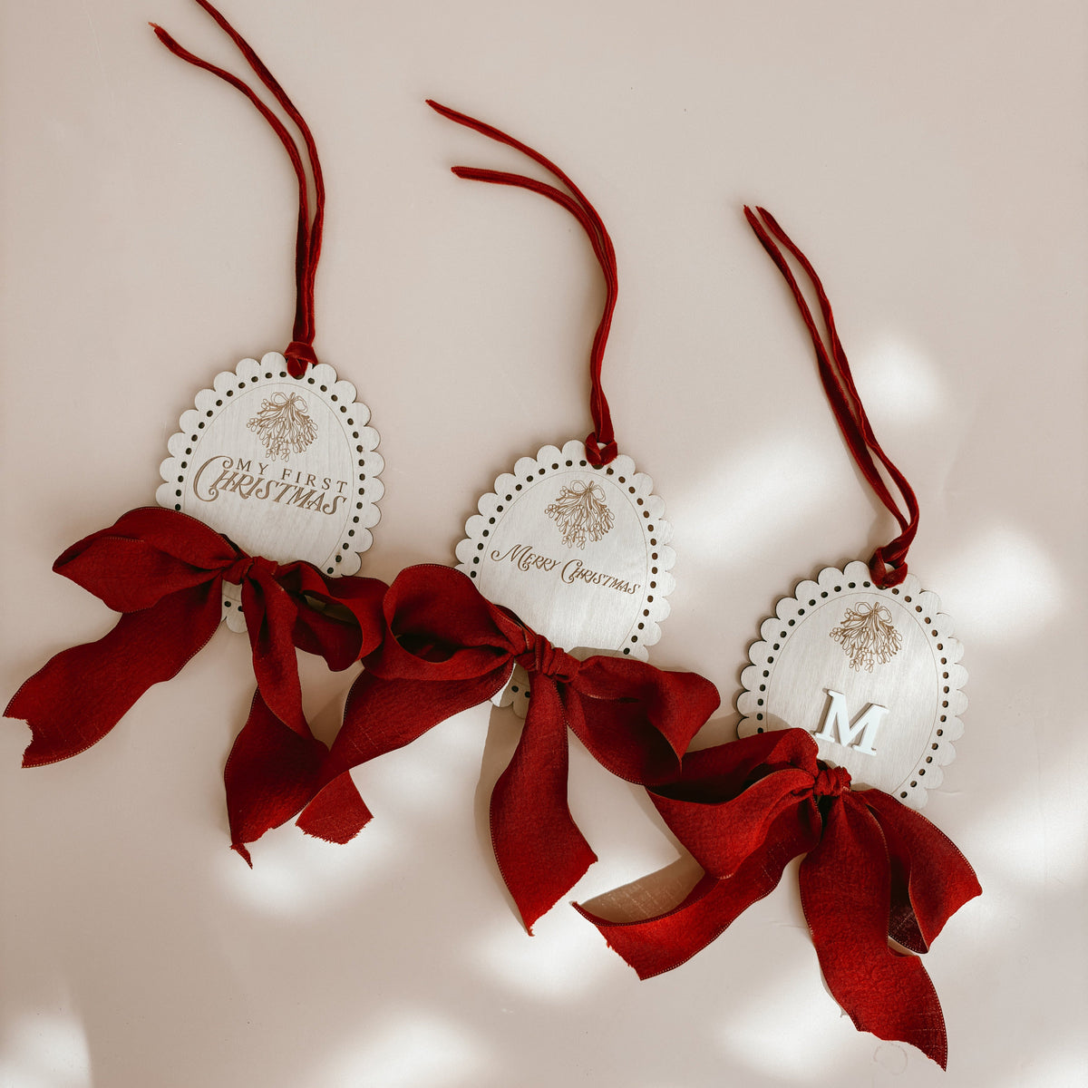 Three oval white Christmas ornaments with scalloped edges, hanging red ribbon bows, and red hanging cords, arranged on a light beige background with soft shadow patterns.