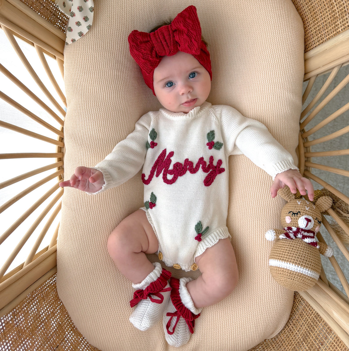 Baby in a cream Merry knit romper and red knot headband lying in a rattan bassinet with knit booties and a crochet reindeer rattle
