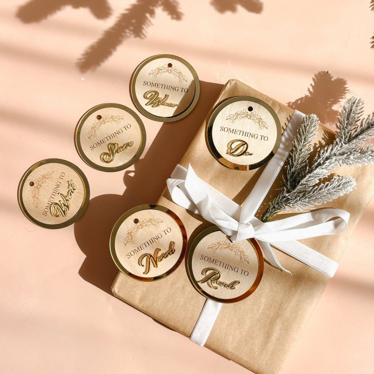 A styled flat-lay of round kraft gift tags with gold and white borders and cursive text, arranged around a kraft-wrapped gift box tied with a white ribbon on a peach background with pine branches and shadows.