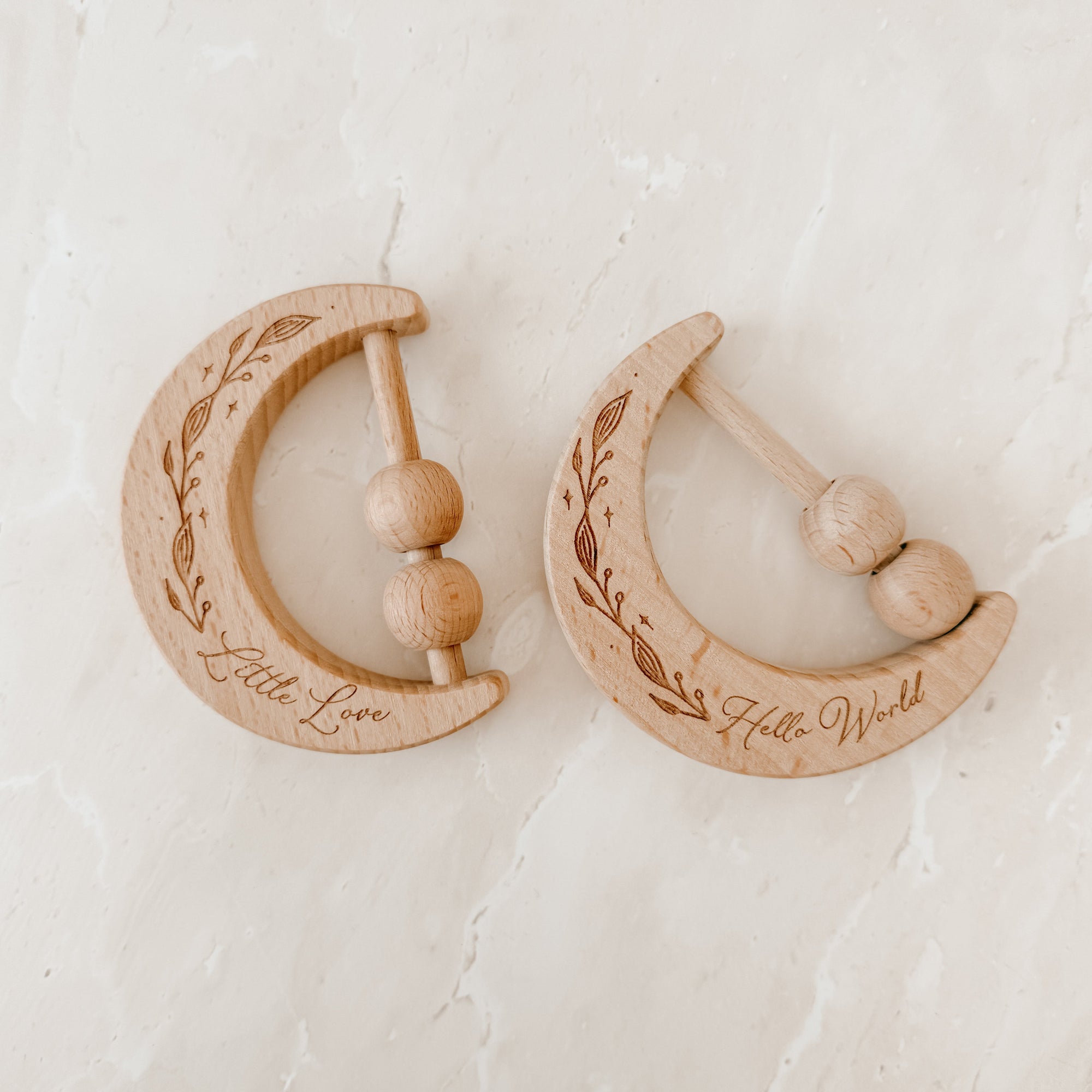Pair of wooden crescent moon shaped baby rattles with engraved 'Little Love" and "Hello World" text on a textured beige surface