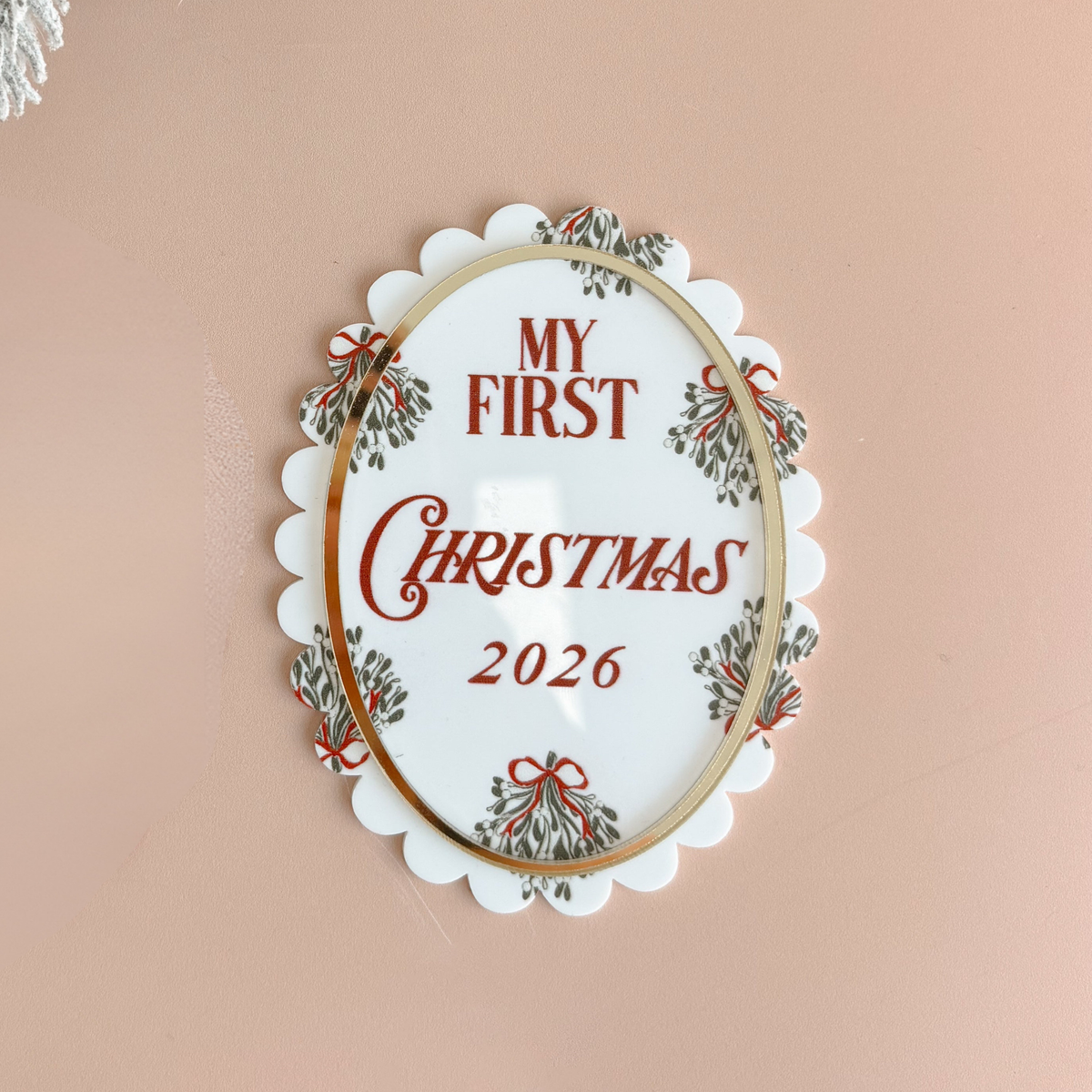 An oval white ceramic Christmas ornament with scalloped edges, featuring the text 'MY FIRST CHRISTMAS 2026' in red lettering, surrounded by a gold inner border and green holly leaf motifs with red bows, set against a solid dusty pink background.
