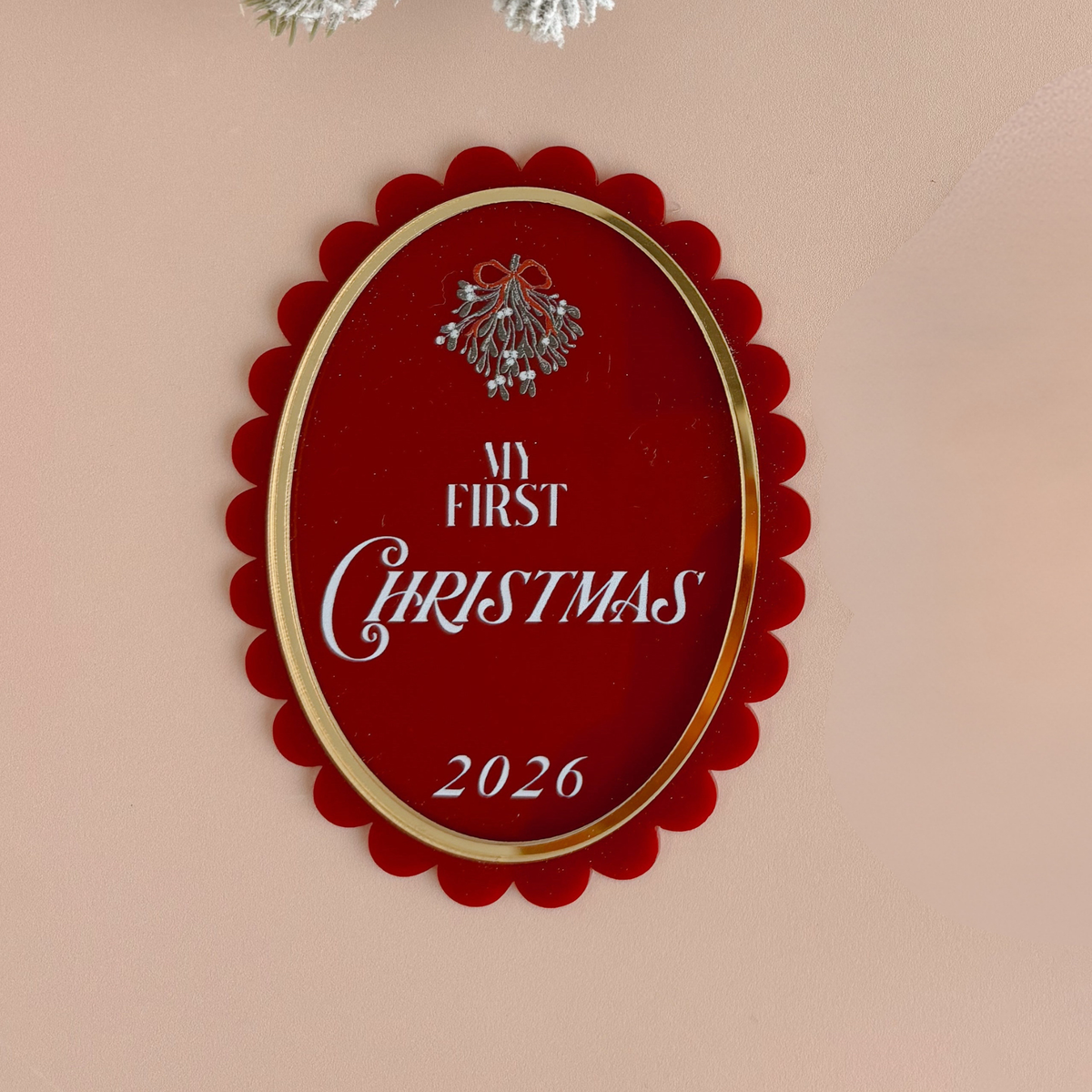 An oval red Christmas ornament with a gold scalloped border and a silver mistletoe graphic, featuring the text 'MY FIRST CHRISTMAS 2026' in white, displayed on a beige wall.