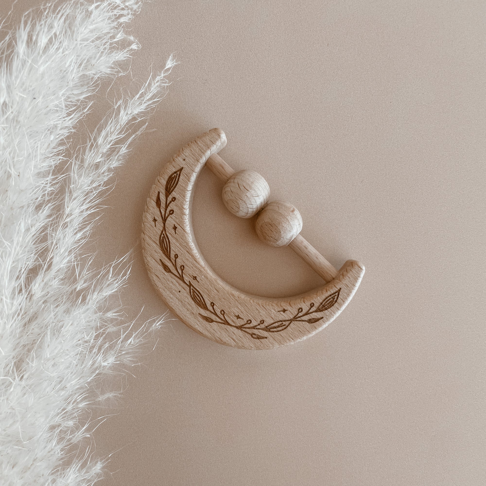 Wooden baby rattle shaped like a crescent moon with engraved patterns on a beige background.