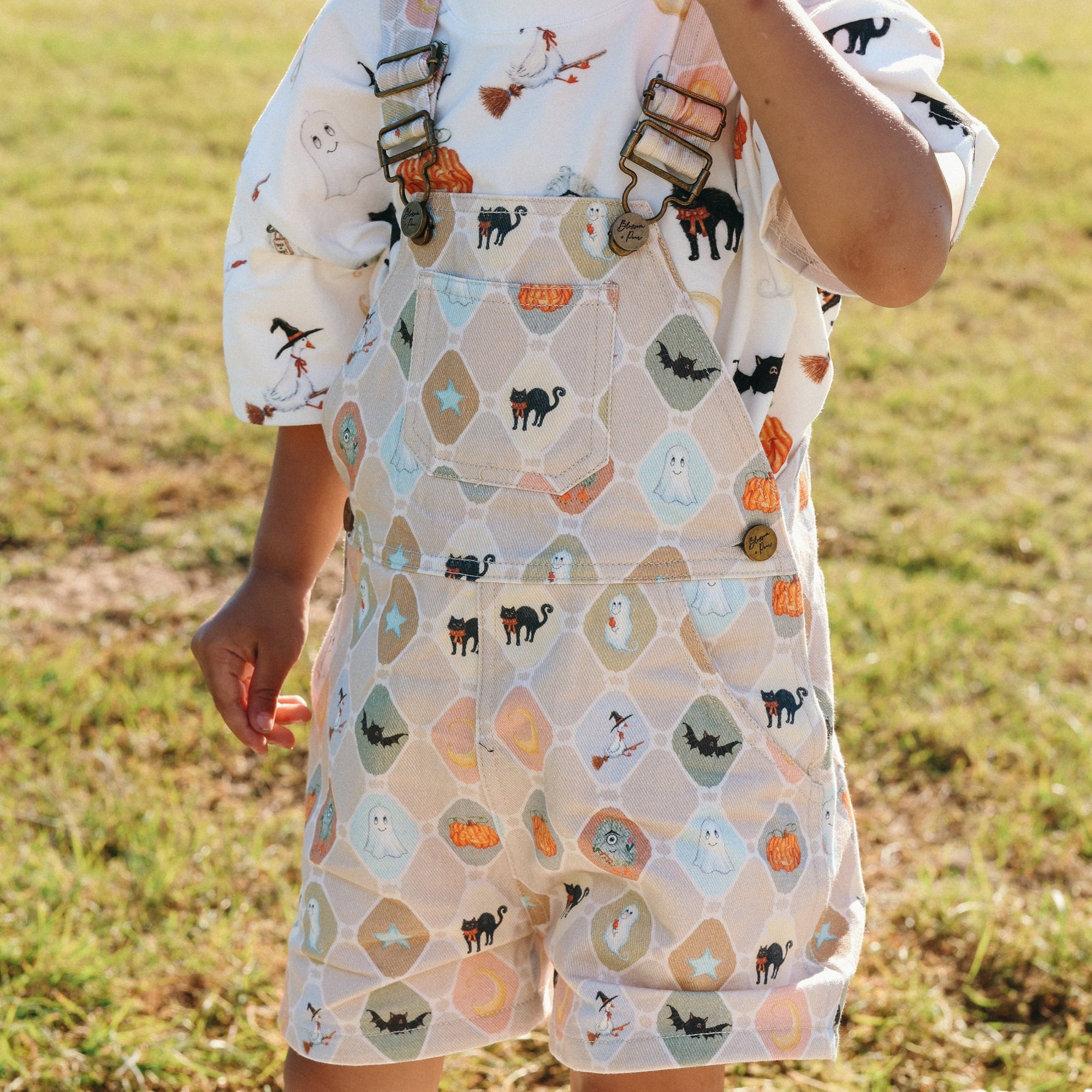 Close up of Halloween print overalls with ghost, bat and pumpkin tiles worn over a matching print shirt