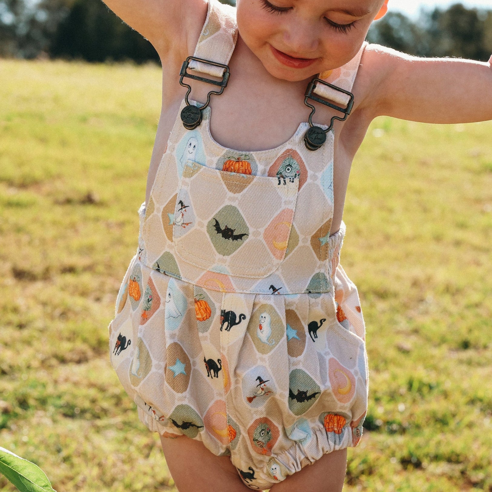 Toddler in a Halloween print bubble overall romper reaching up, arms out, in a sunlit field