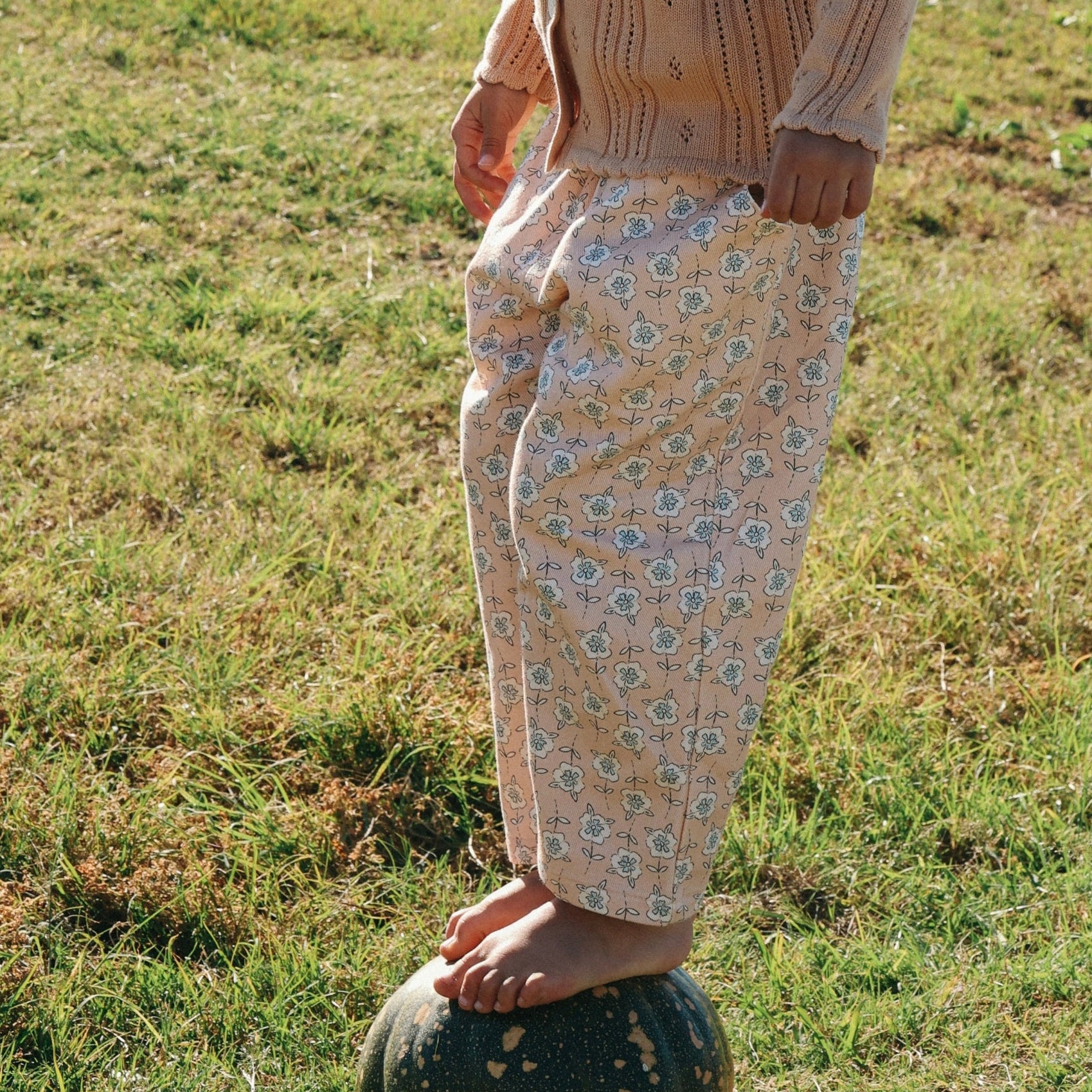 Child in a cream pointelle knit top and floral print pants standing on a pumpkin in a sunlit field