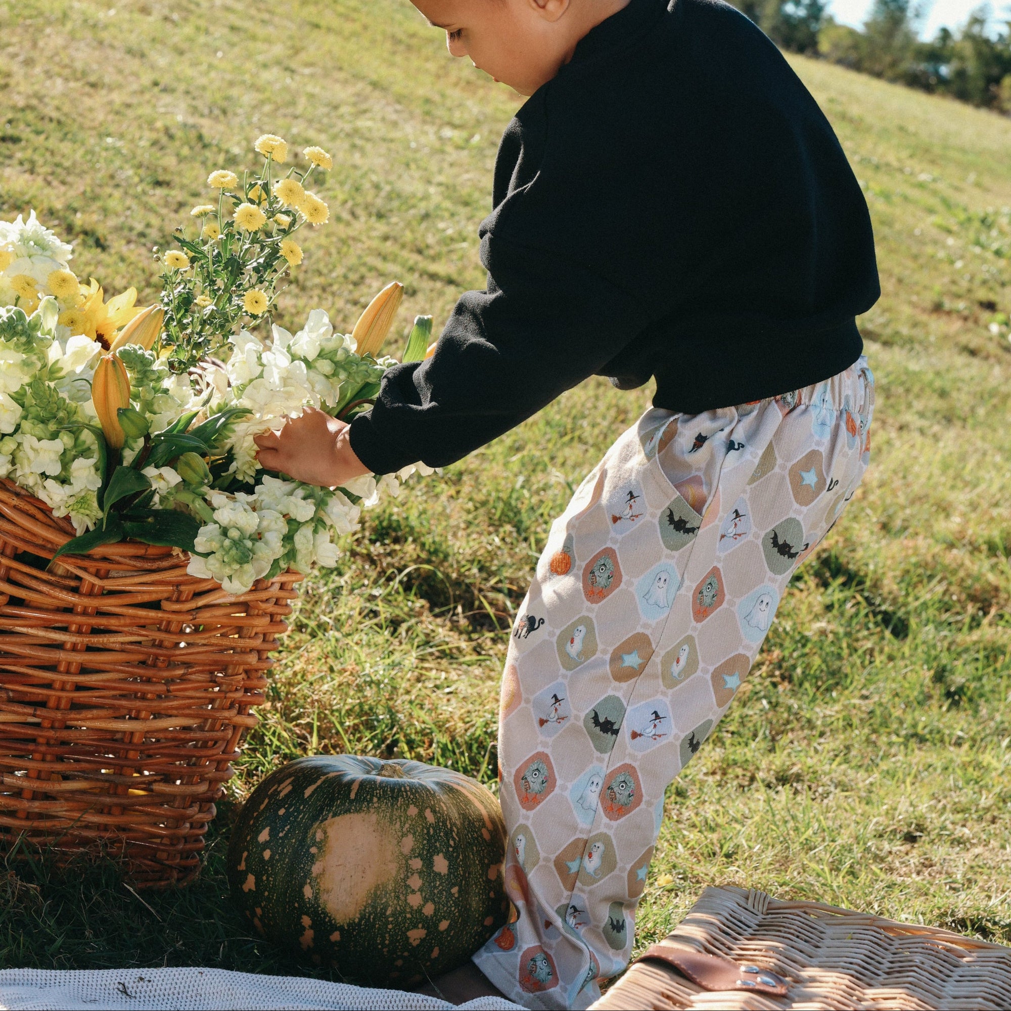 Toddler in a black sweatshirt and Halloween print pants picking flowers from a basket beside a pumpkin