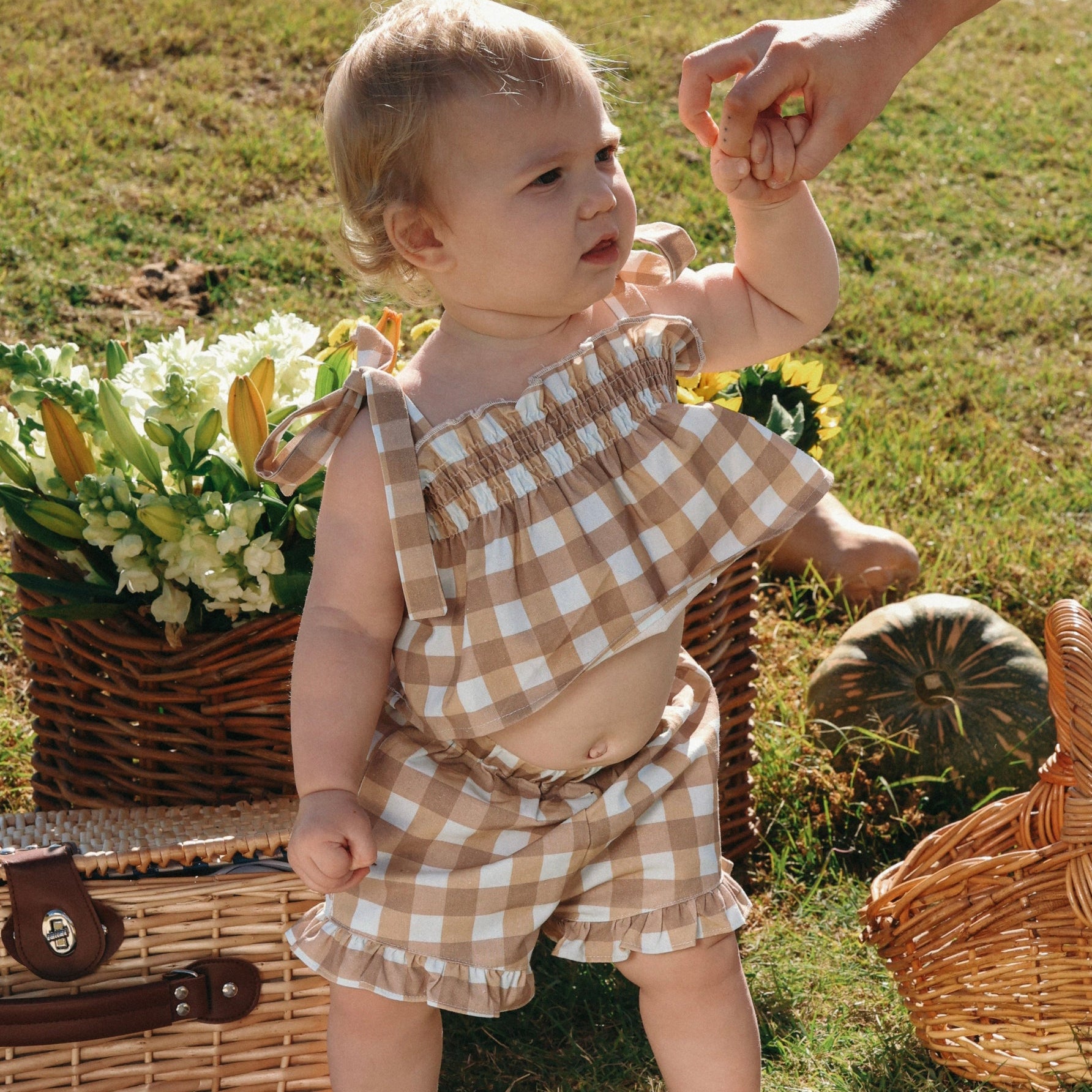 Baby in a tan gingham shirred top and ruffle shorts set standing beside flower baskets and pumpkins