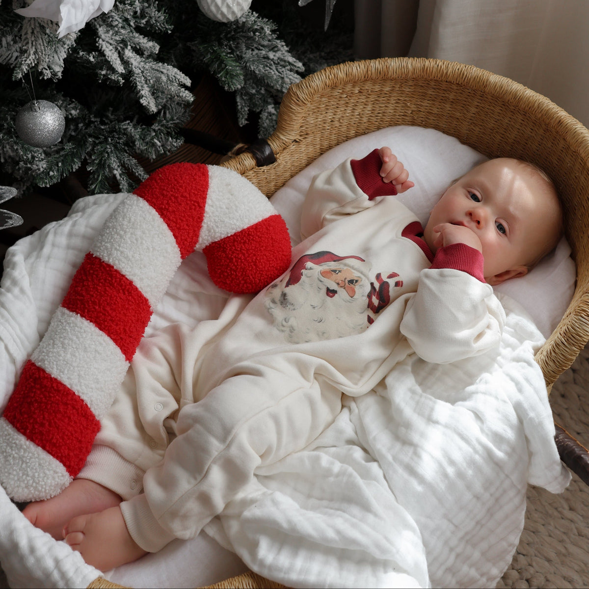 A baby wearing a white Christmas-themed onesie lies in a woven bassinet next to a decorated Christmas tree, with a red and white striped stocking beside them.