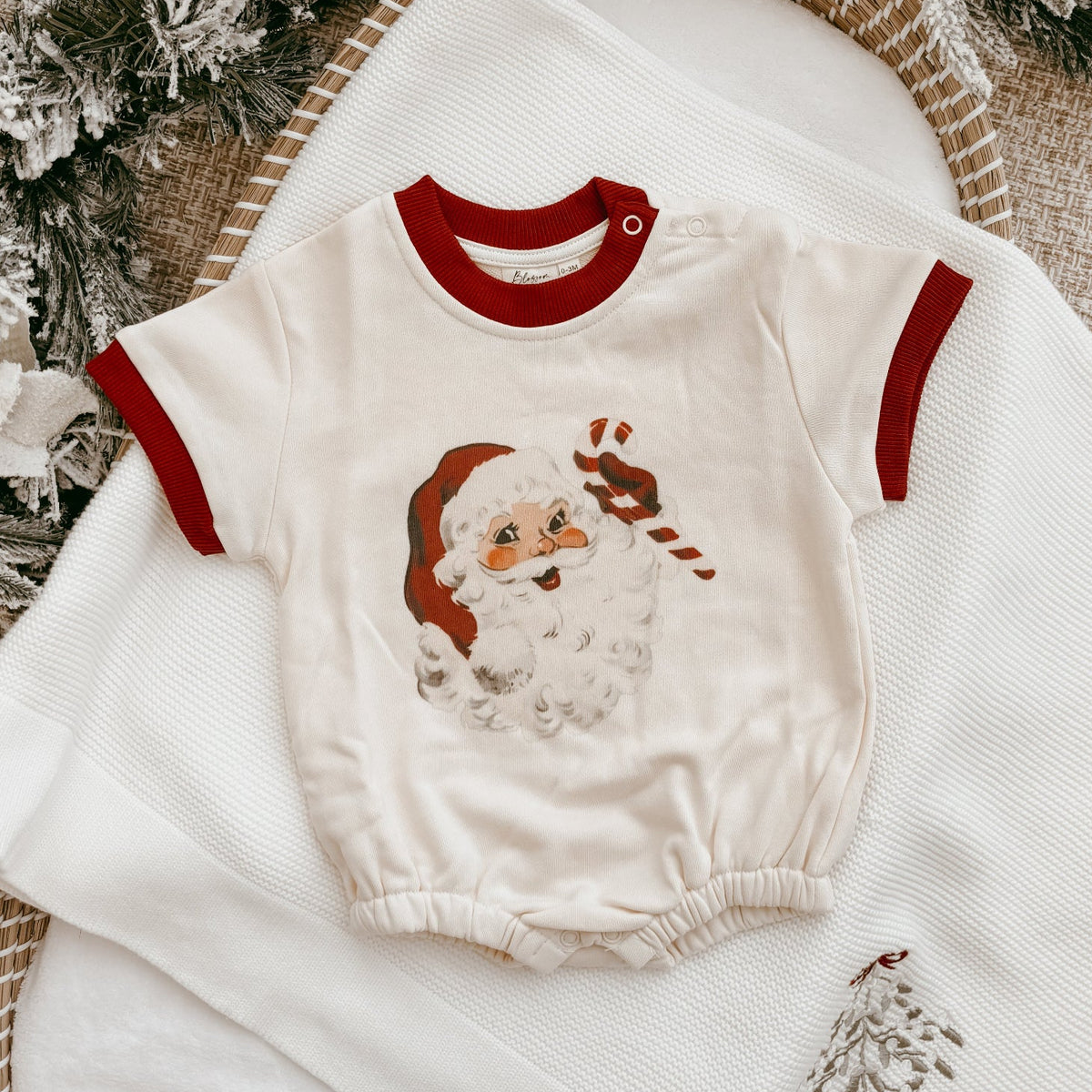 A baby romper in cream with red trim and a Santa Claus graphic, styled on a white blanket with holiday garlands and a woven mat.