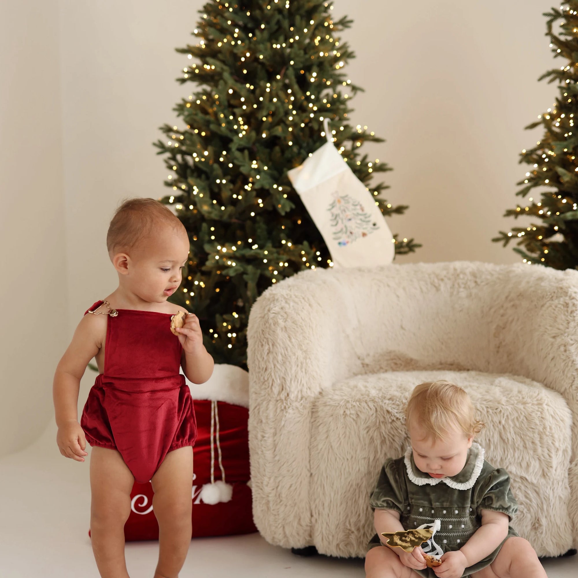 Two babies in red and green velvet Christmas rompers eating cookies beside a lit Christmas tree