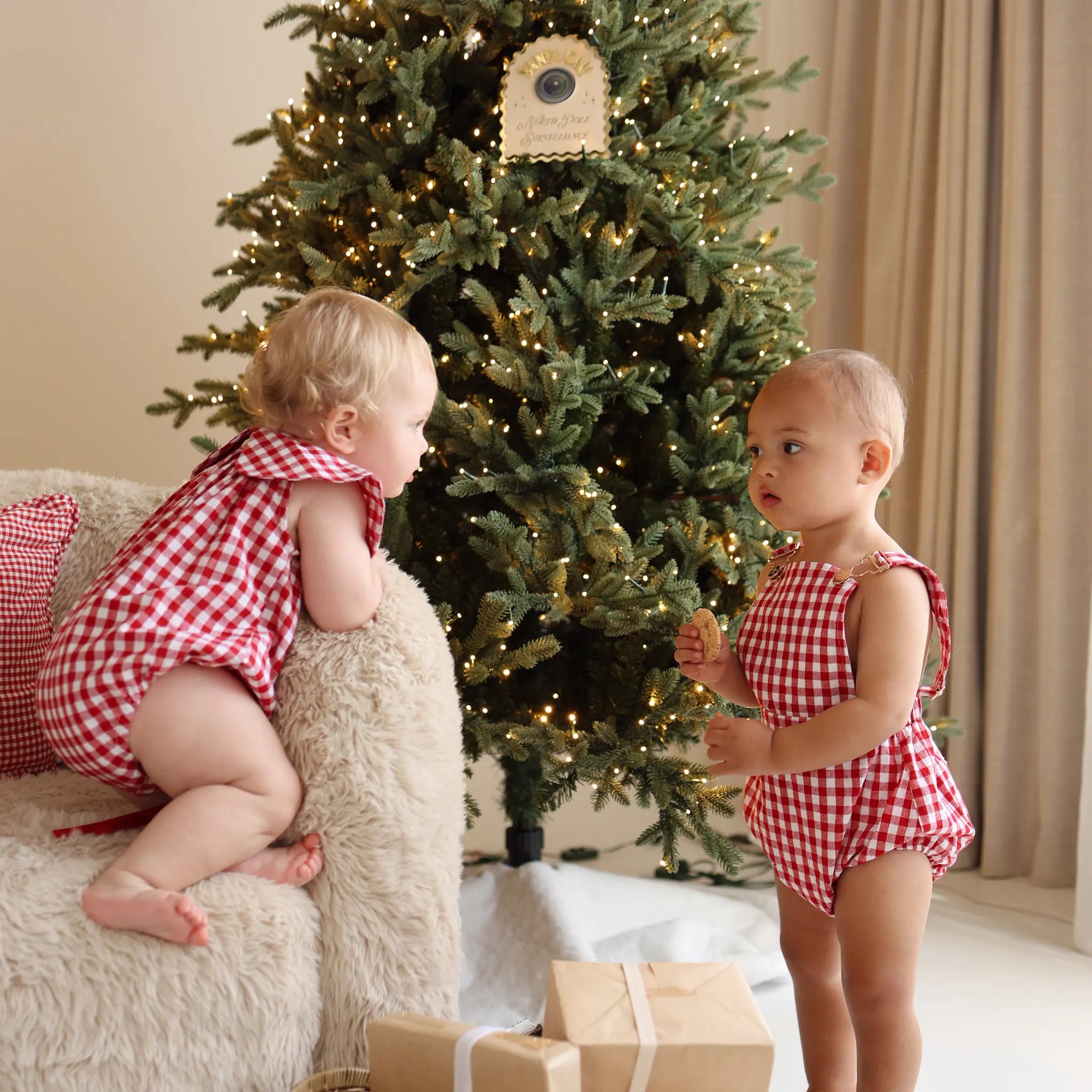 Two babies in matching red gingham rompers beside a lit Christmas tree and wrapped gifts
