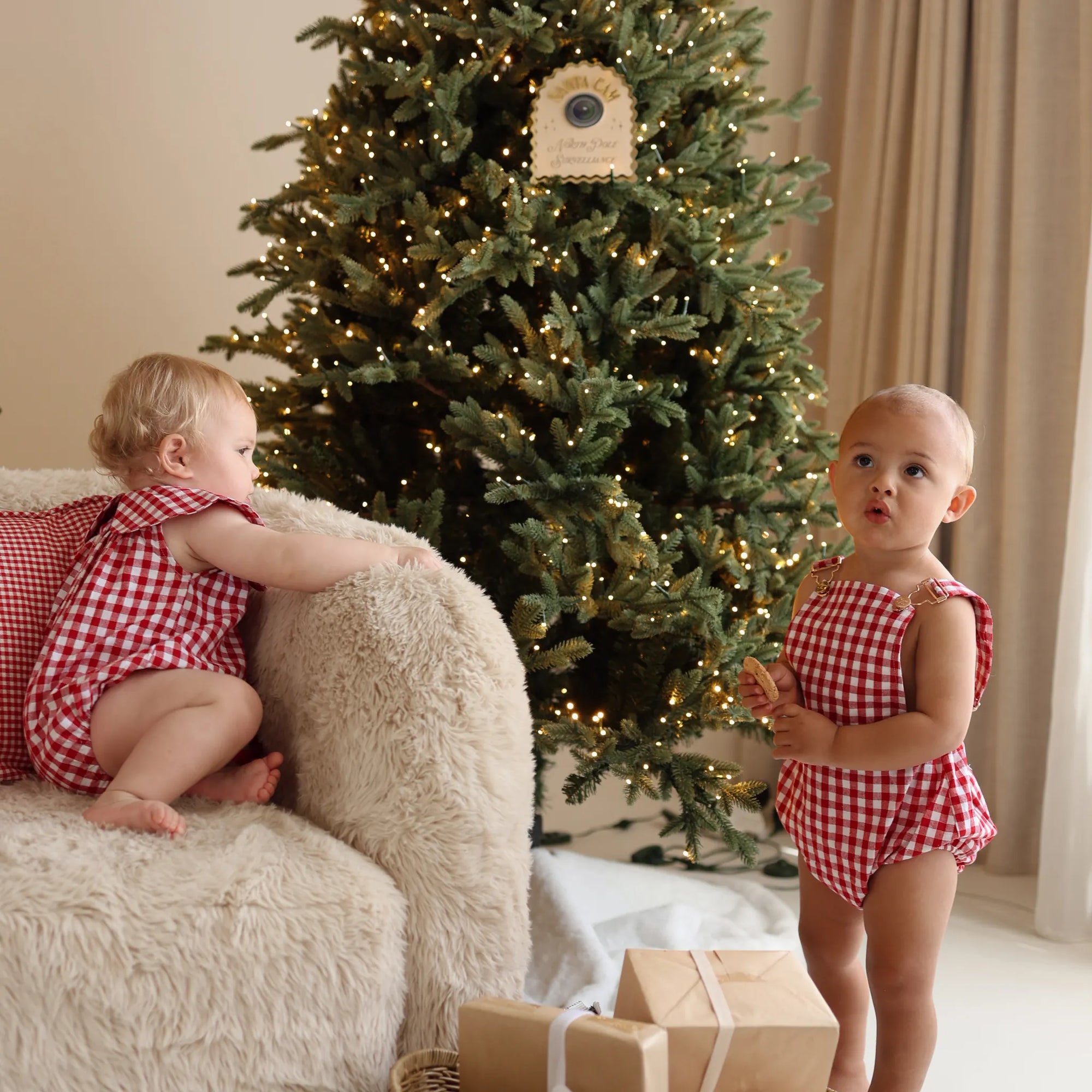 Two babies in red gingham rompers playing by a sheepskin chair and lit Christmas tree