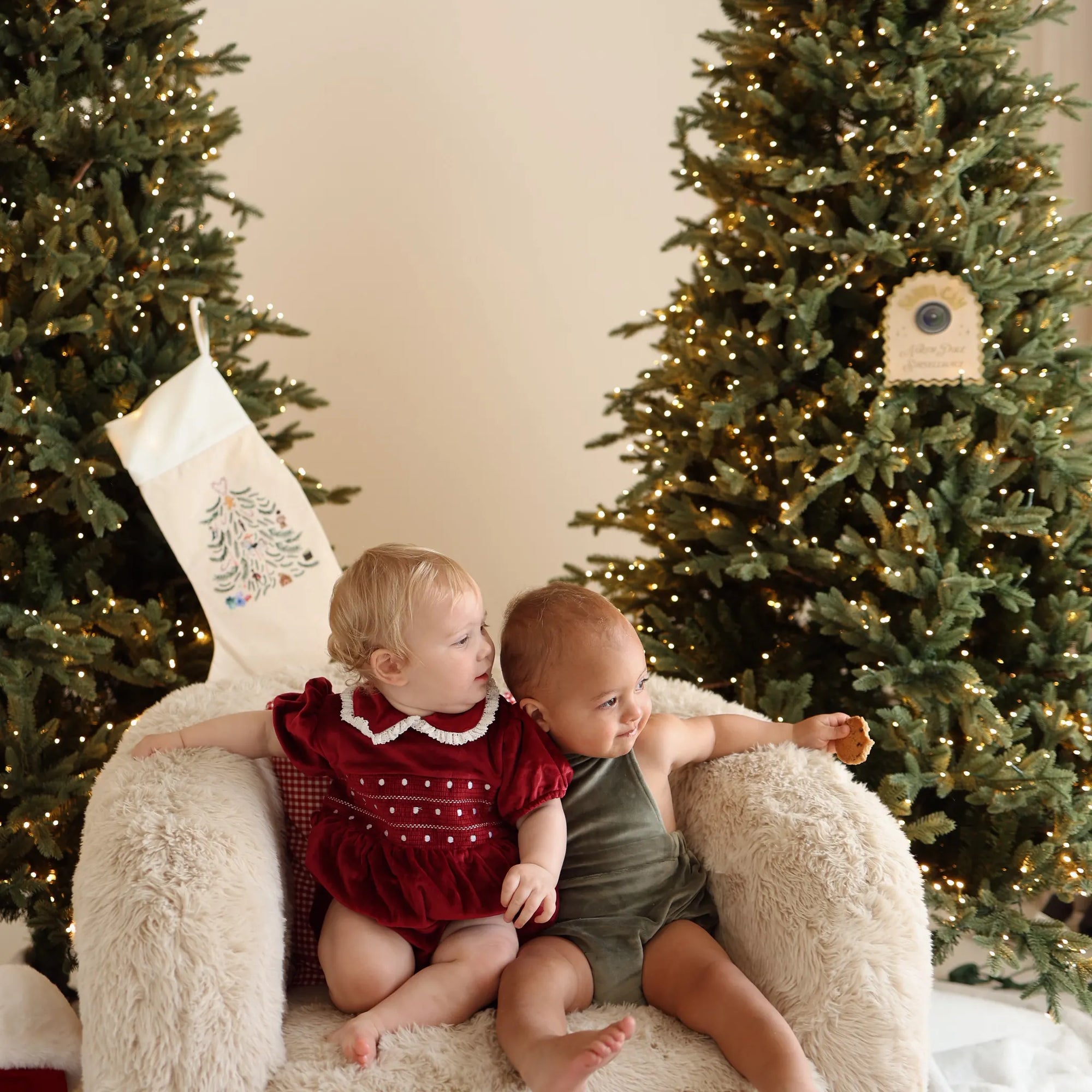 Two babies in red and green velvet Christmas rompers sitting together on a sheepskin chair