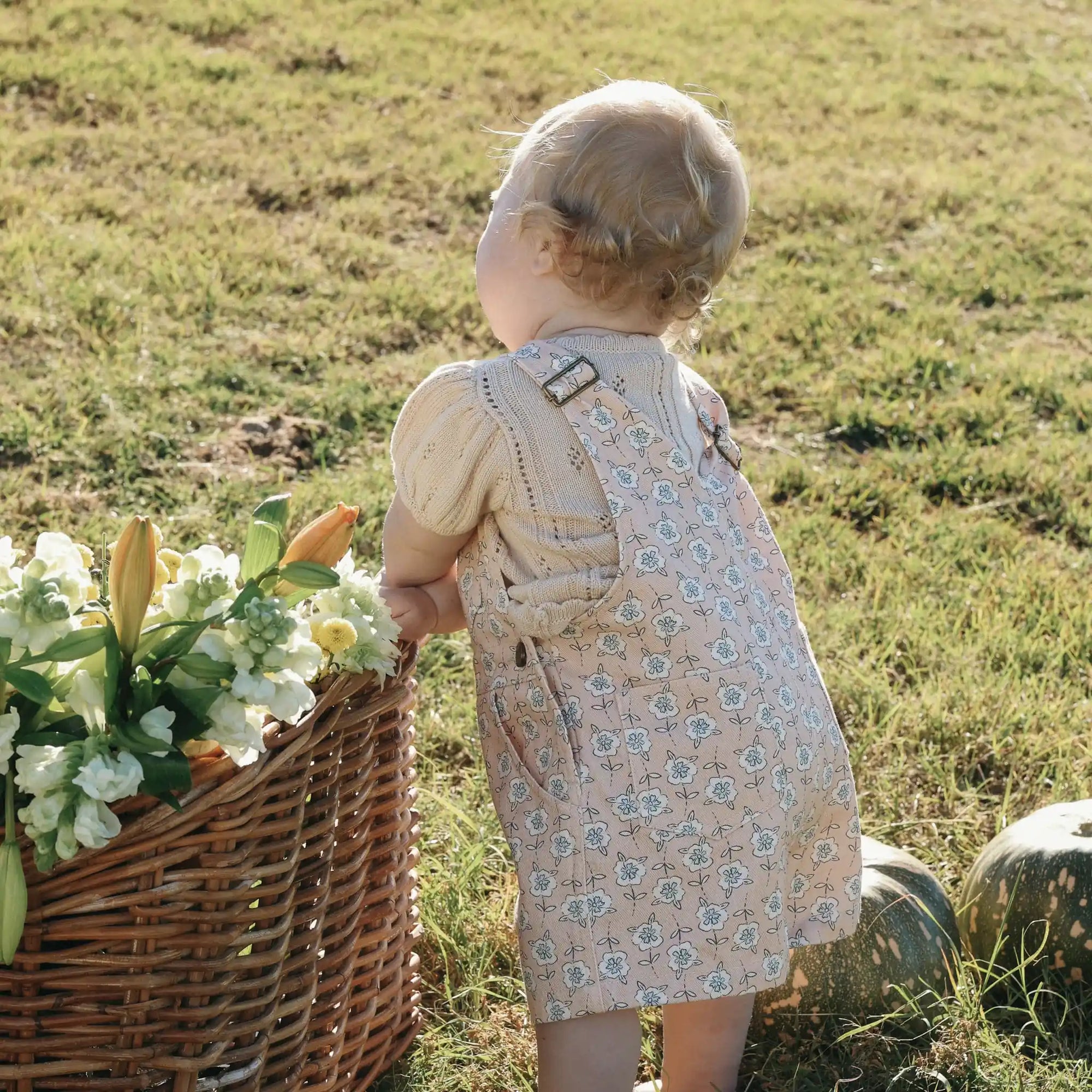 Back detail of floral print baby overalls with buckle straps, worn over a cream pointelle knit top