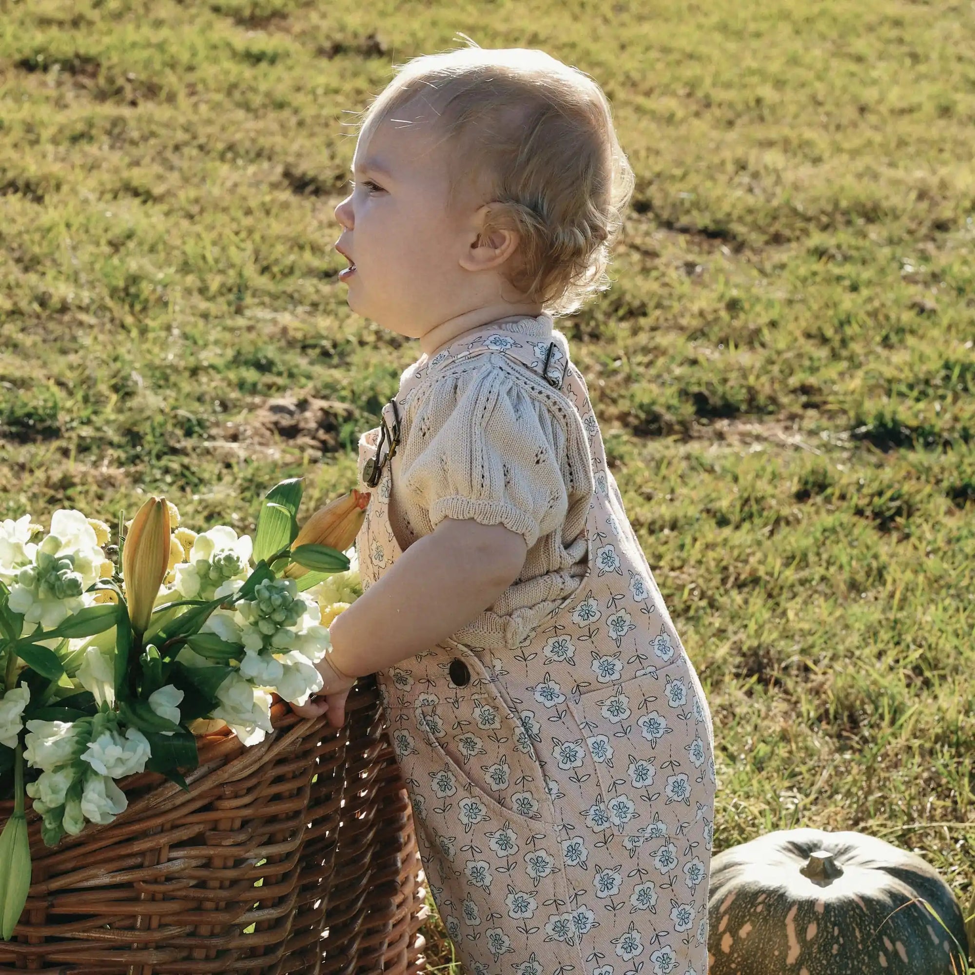 Baby in a cream pointelle top and floral print overalls standing beside a basket of white flowers