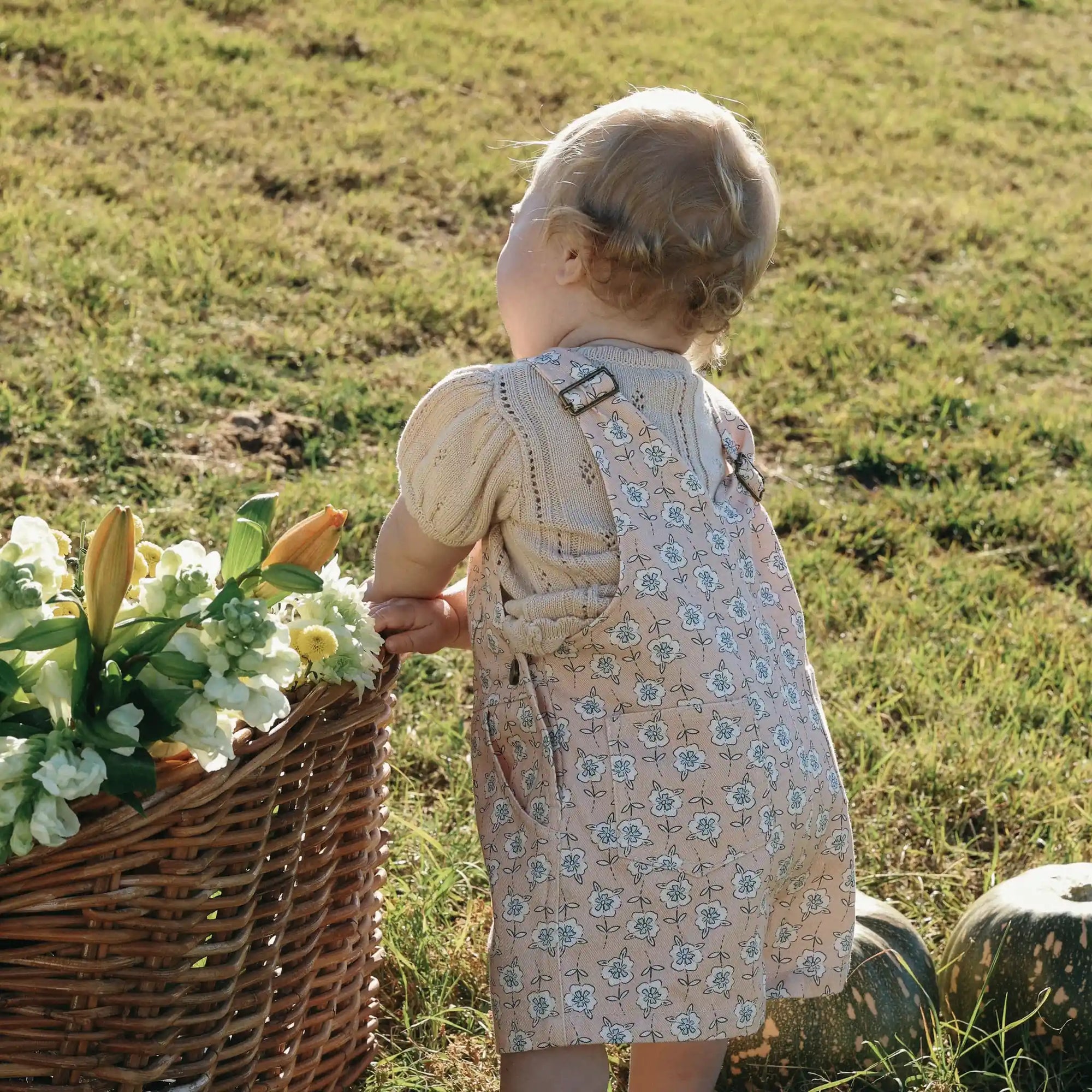 Back view of a baby in floral print overalls and a cream pointelle top beside a basket of flowers
