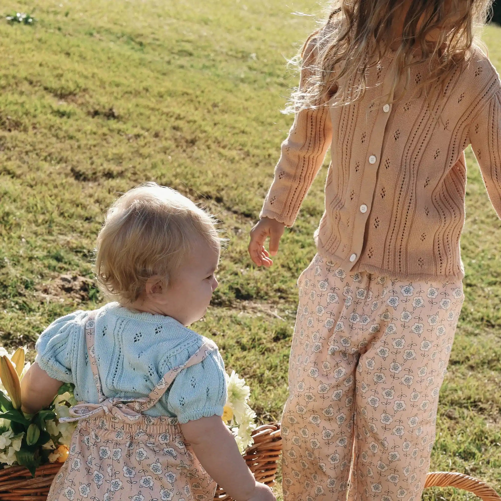 Baby in blue pointelle knit and floral print overalls beside a girl in a matching floral print set