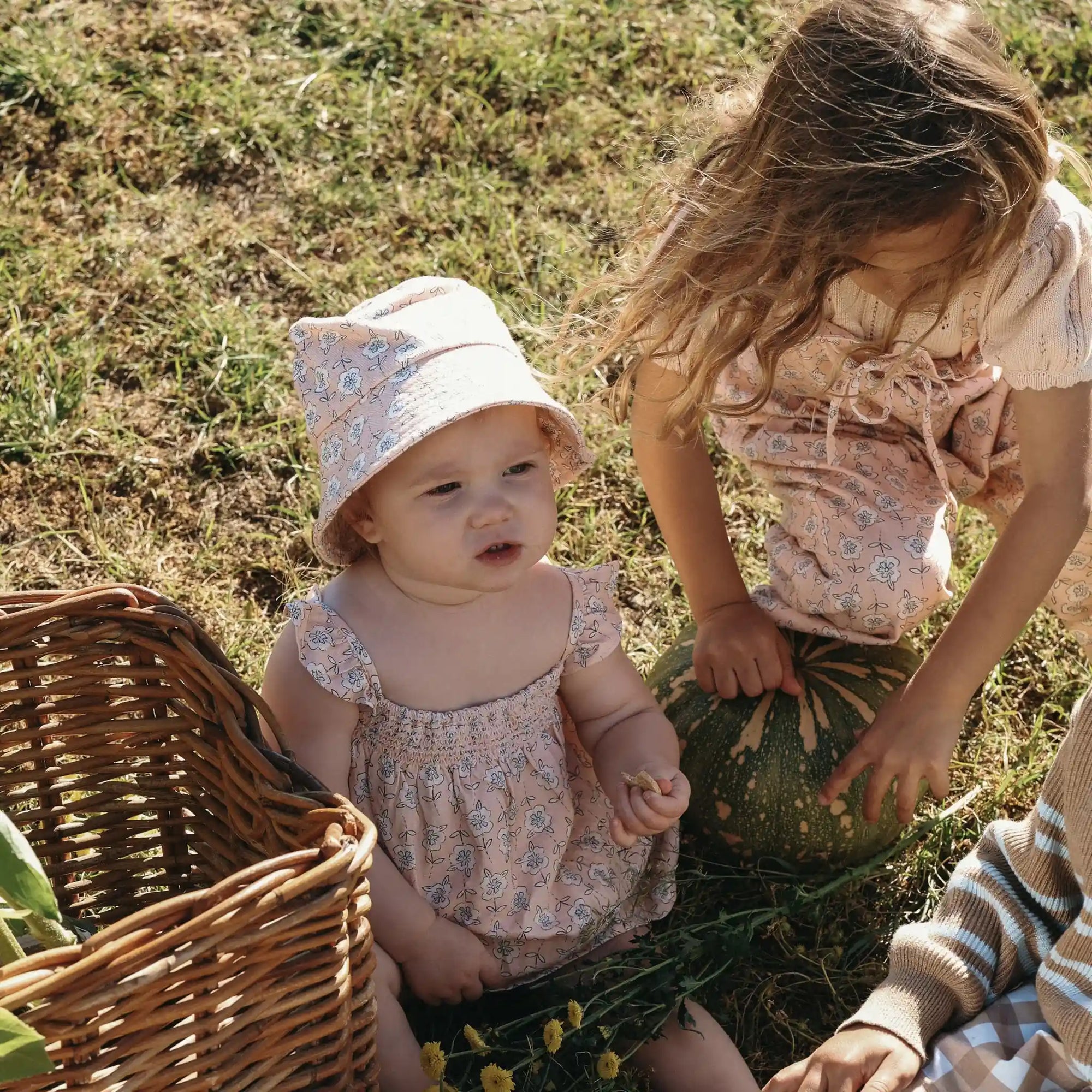 Baby in a floral print romper and bucket hat beside a girl in matching print holding a pumpkin