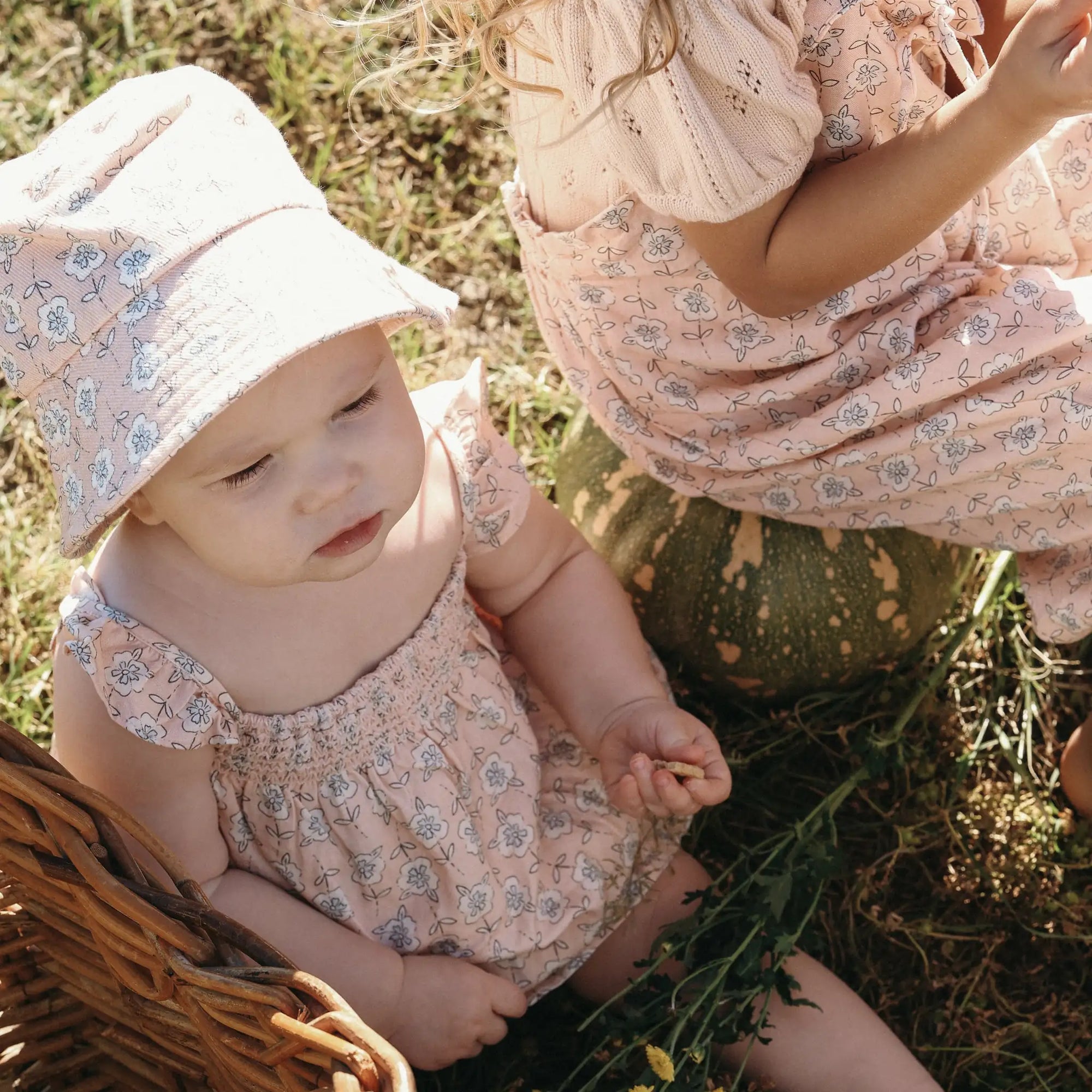 Baby in a floral print romper and bucket hat sitting beside a pumpkin and an older girl in matching print