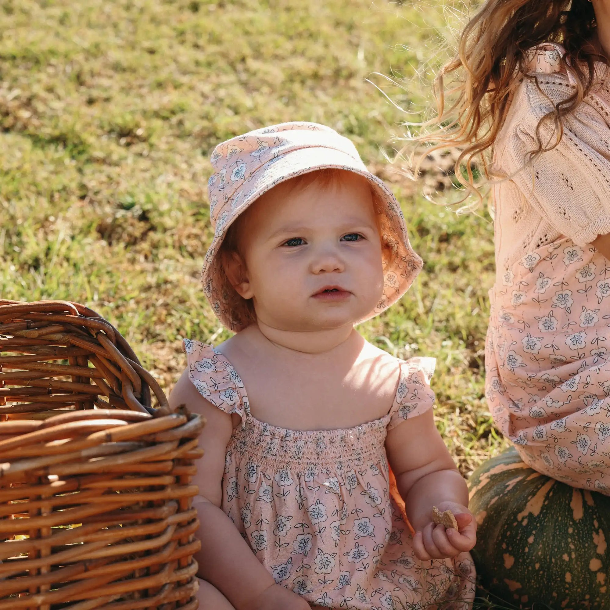 Baby in a floral print shirred romper and matching bucket hat sitting beside a wicker basket