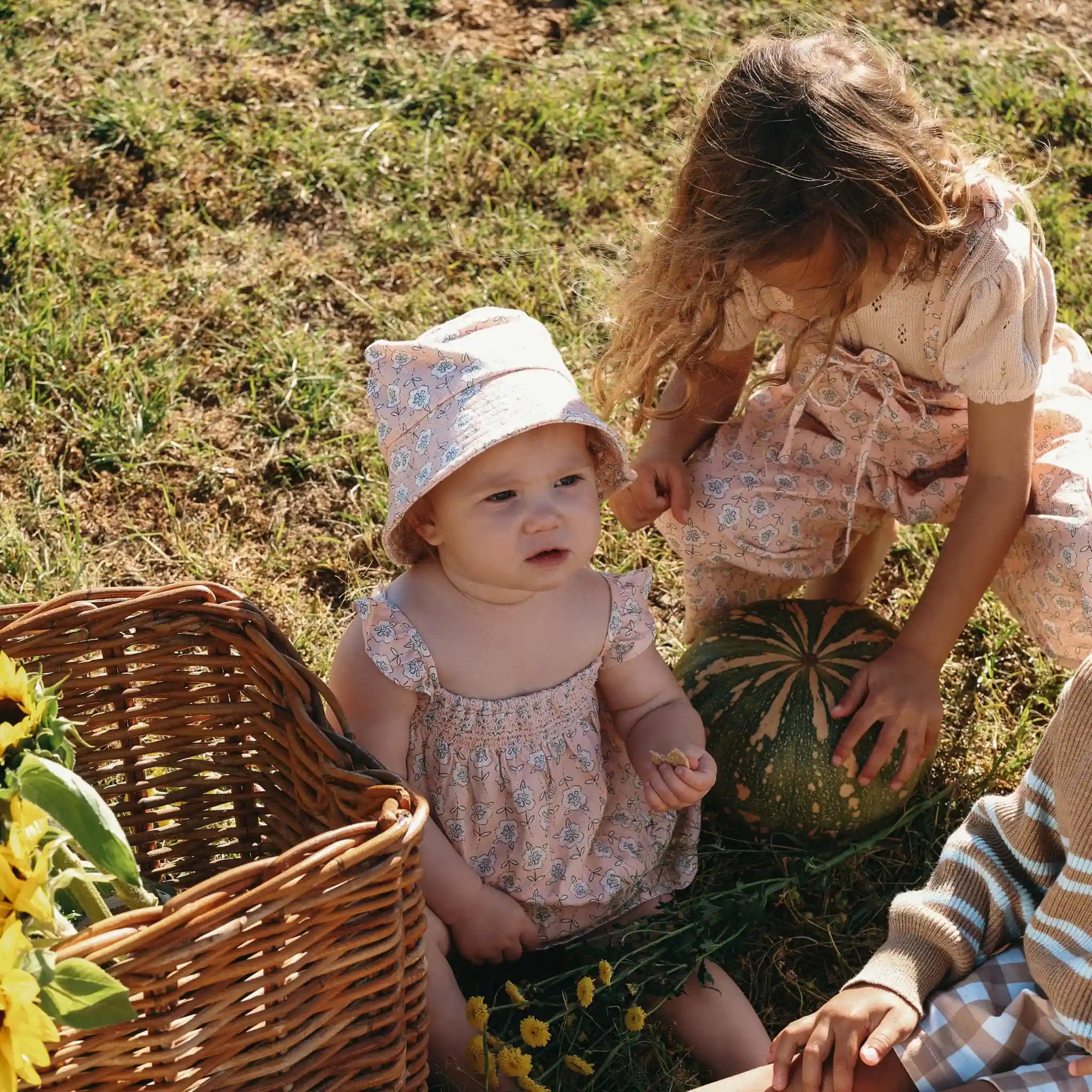 Baby in a floral print romper and bucket hat sitting beside an older sister lifting a pumpkin