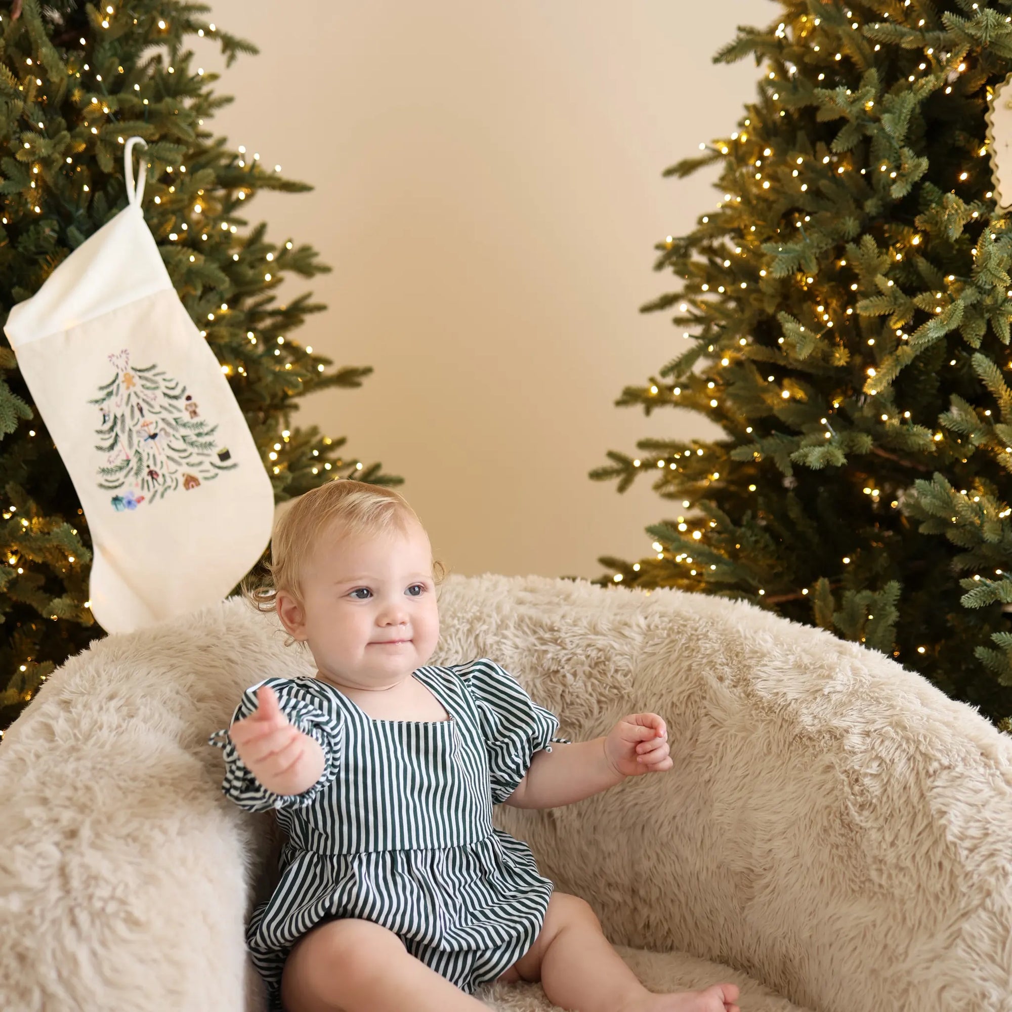 Baby in a green stripe puff sleeve romper sitting on a sheepskin chair beside an embroidered Christmas stocking