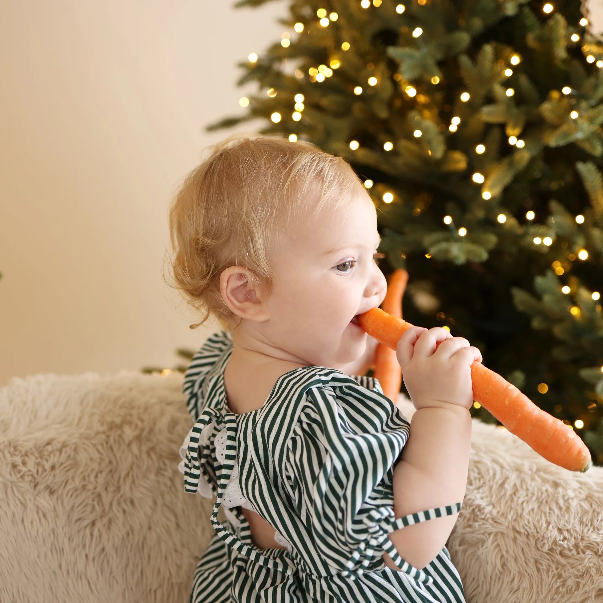 Baby in a green stripe romper biting a carrot left out for the reindeer, beside a lit Christmas tree
