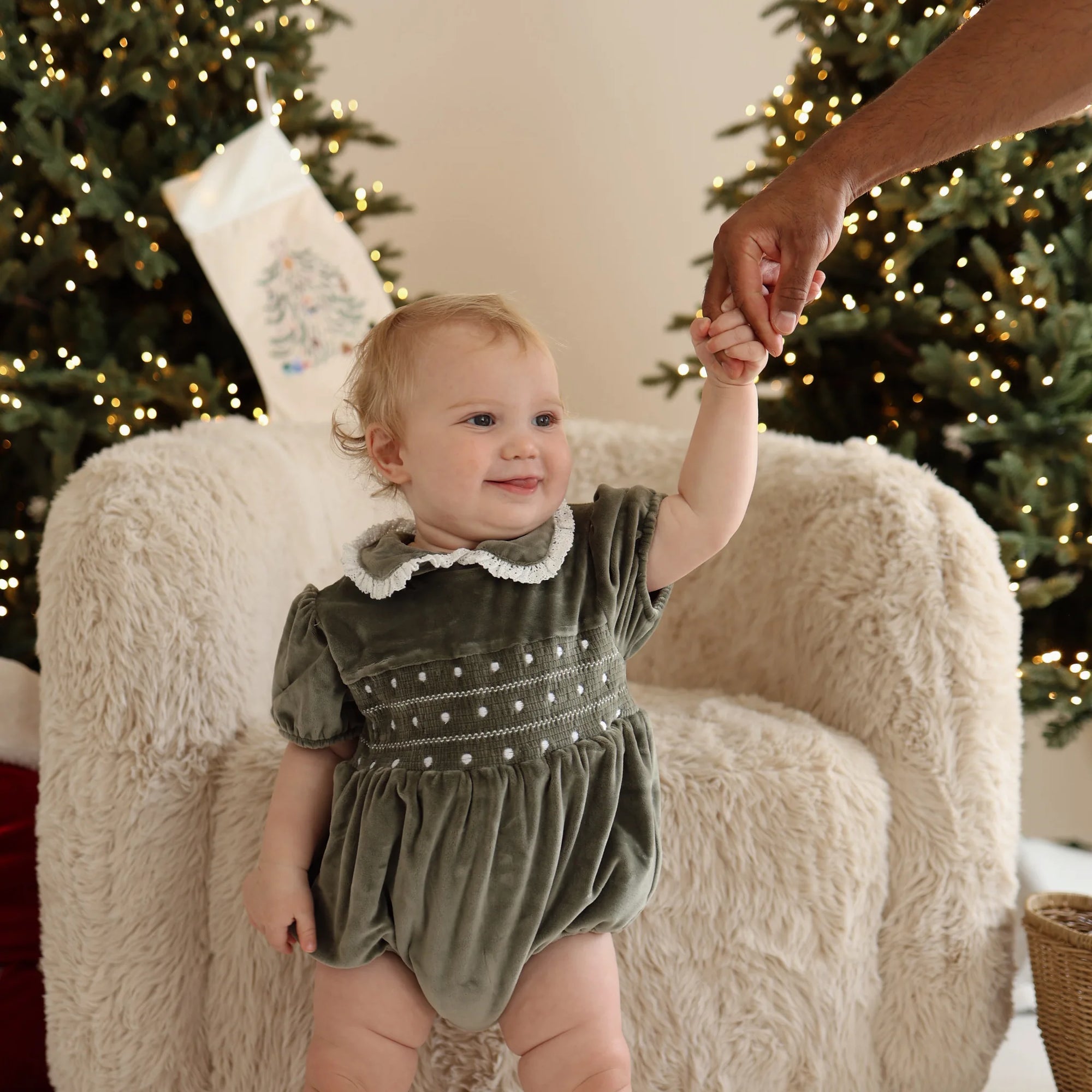 Smiling baby in a green velvet smocked Christmas romper standing by a sheepskin chair