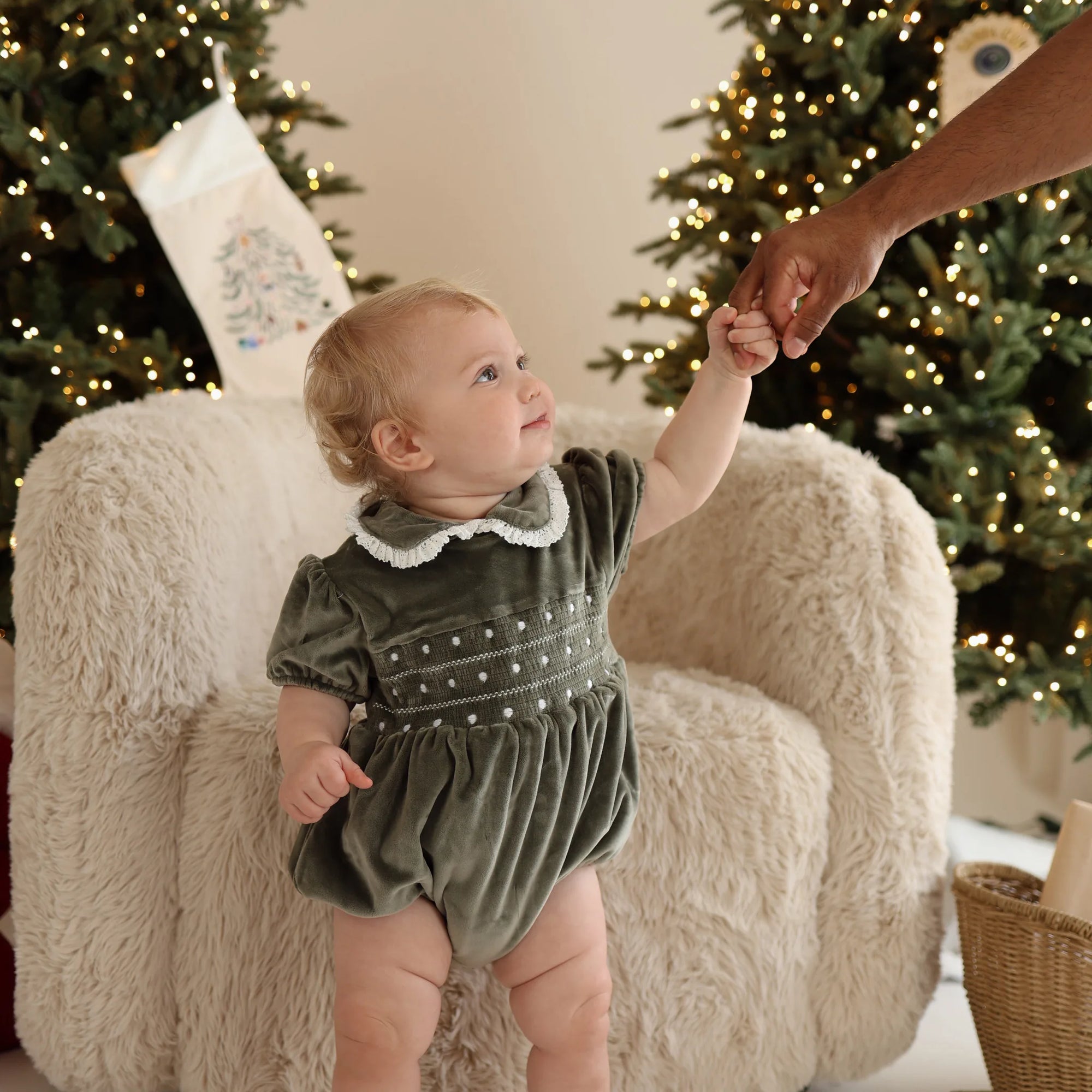 Baby in a green velvet smocked romper with lace collar holding a hand, lit Christmas trees behind