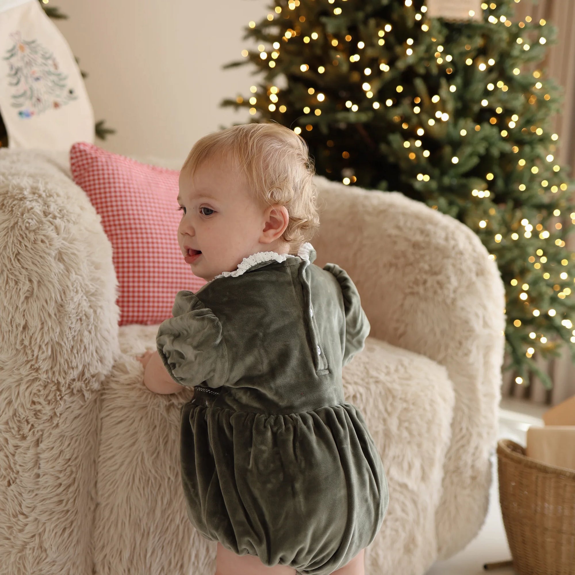 Back view of a baby in a green velvet Christmas romper with lace collar beside a lit tree