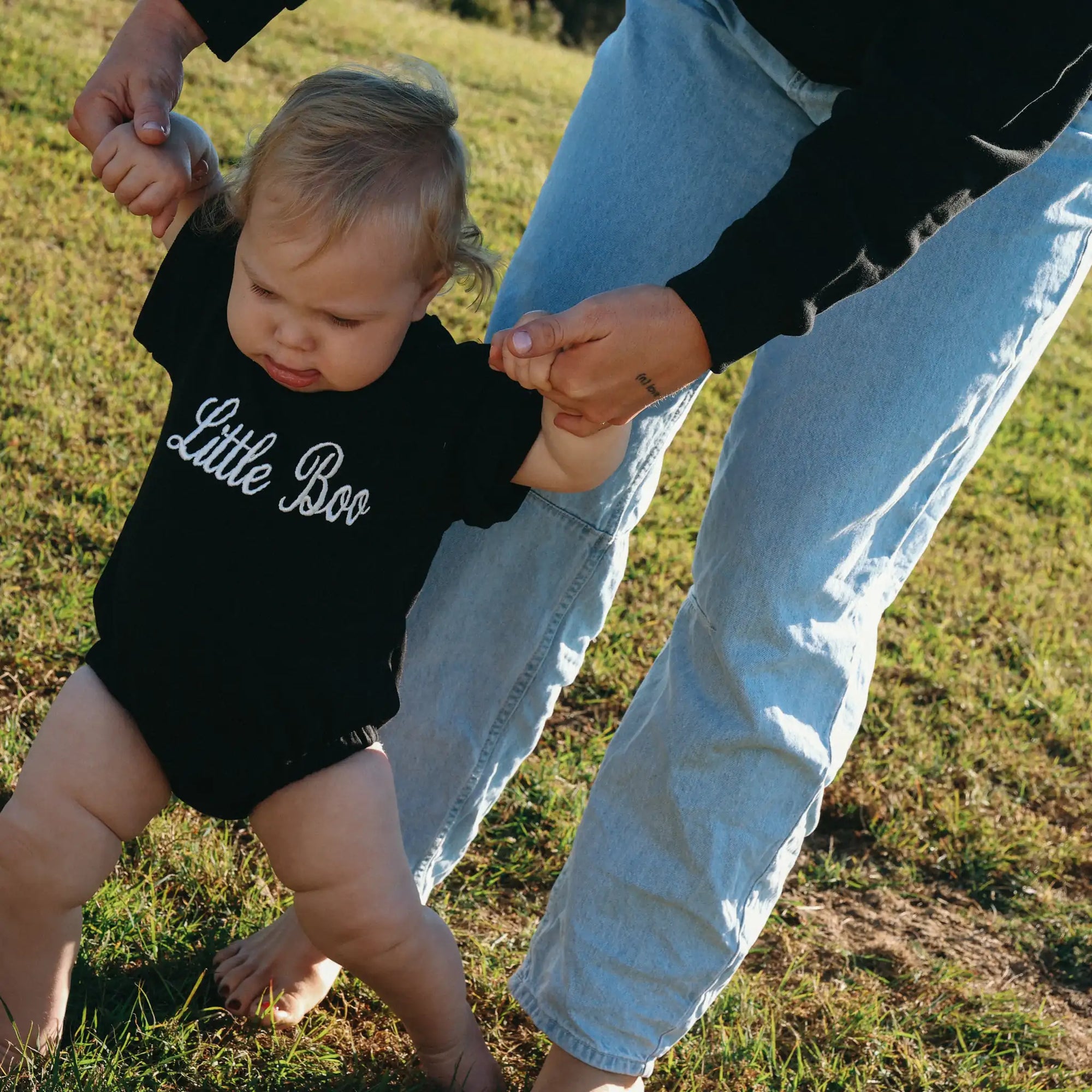Baby in a black Little Boo romper taking first steps holding a parent's hands on grass
