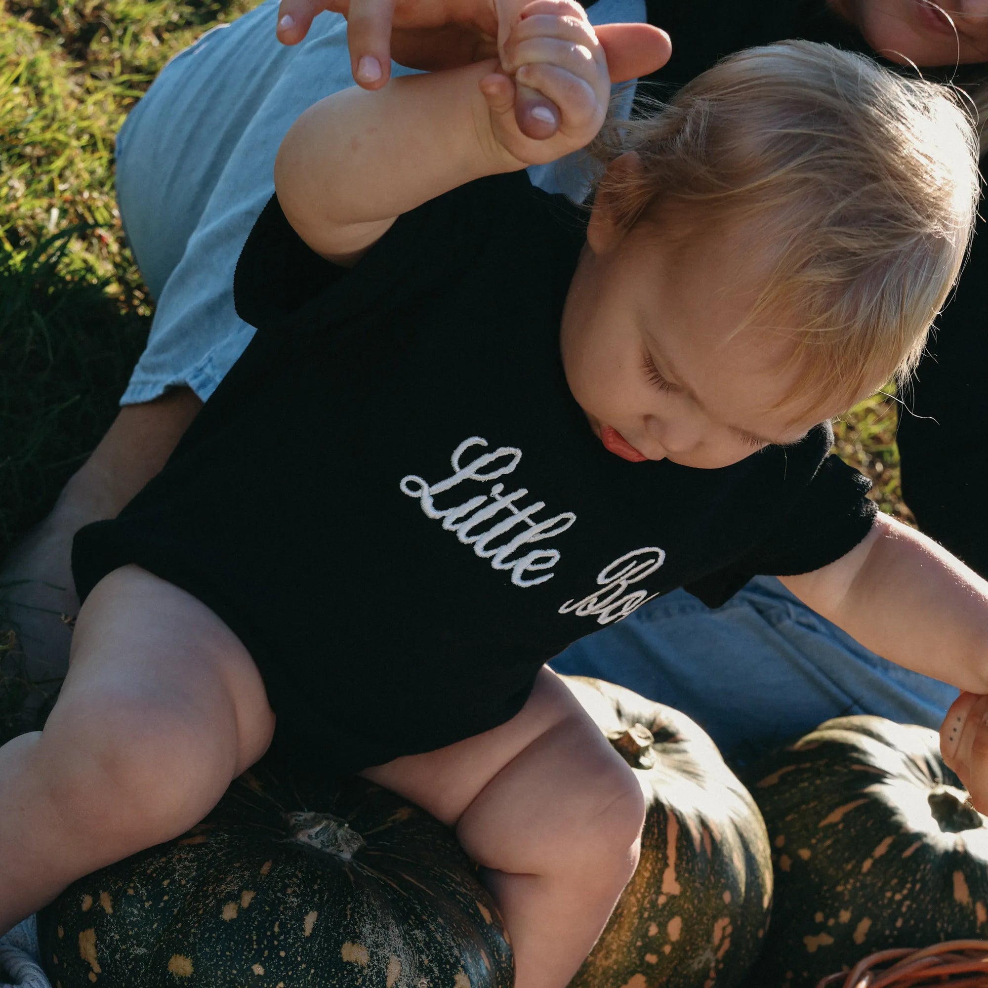 Baby in a black Little Boo embroidered romper being held on a pumpkin in a sunlit field