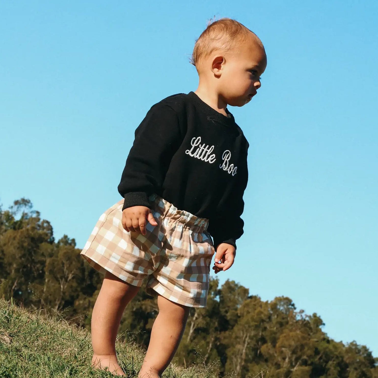 Baby in a black Little Boo sweatshirt and tan gingham shorts walking on a grassy hill against a blue sky