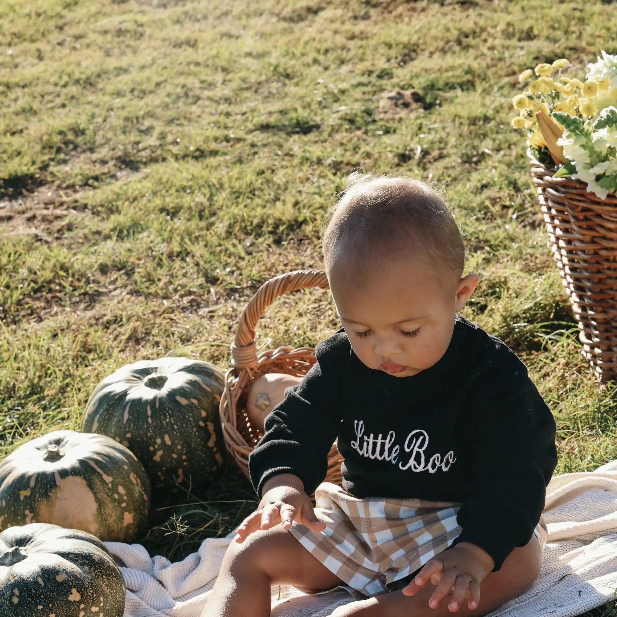 Baby in a black Little Boo embroidered sweatshirt sitting on a blanket beside pumpkins and flowers