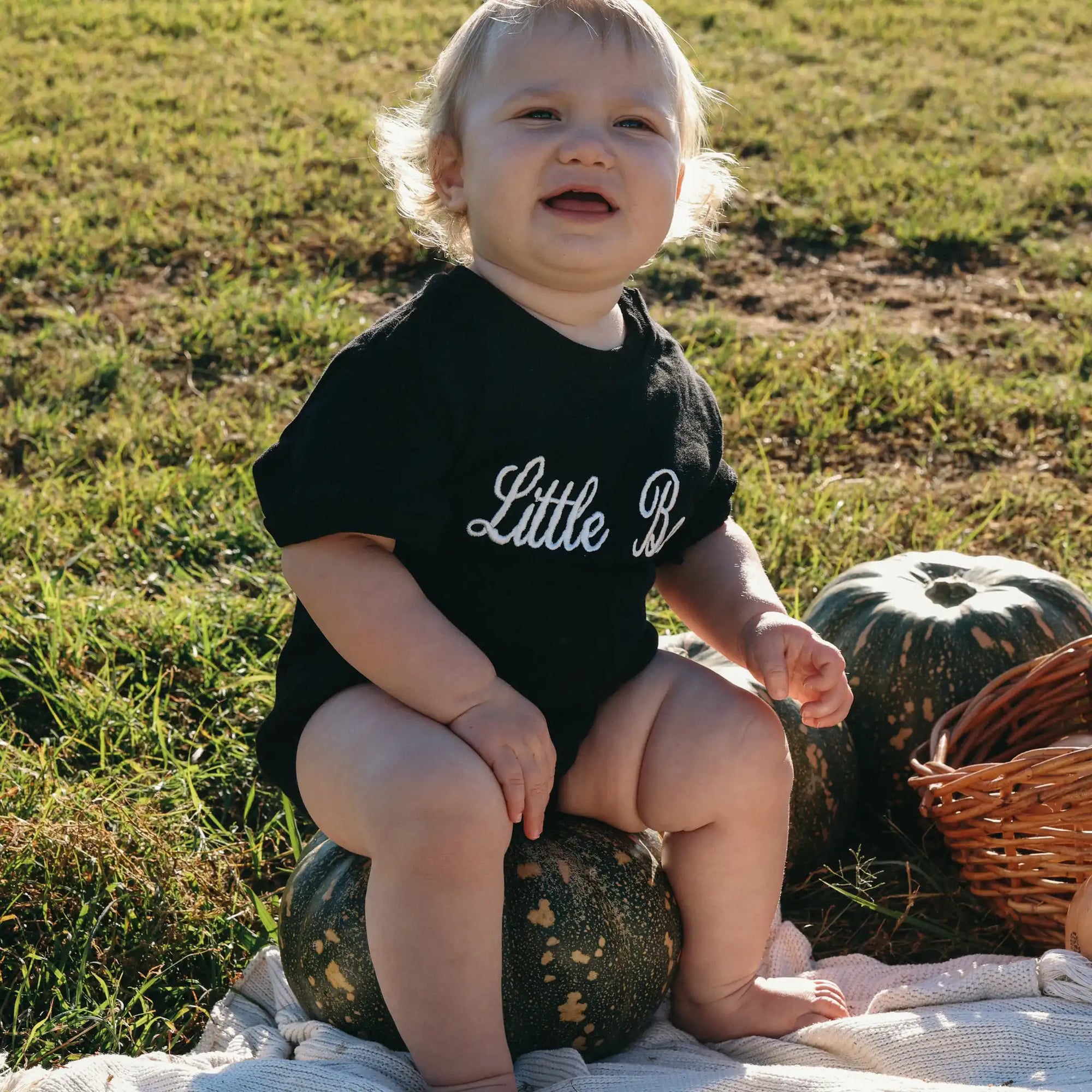 Smiling baby in a black Little Boo embroidered romper sitting on a pumpkin on sunlit grass