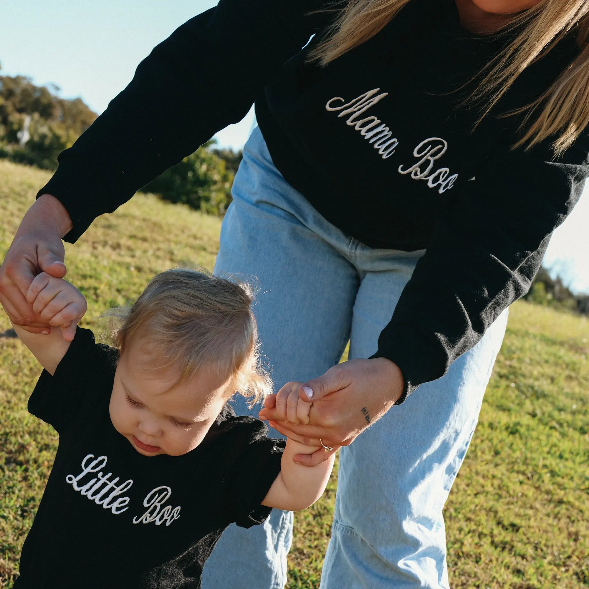Mum in a Mama Boo sweatshirt walking a baby in a matching Little Boo romper across sunlit grass