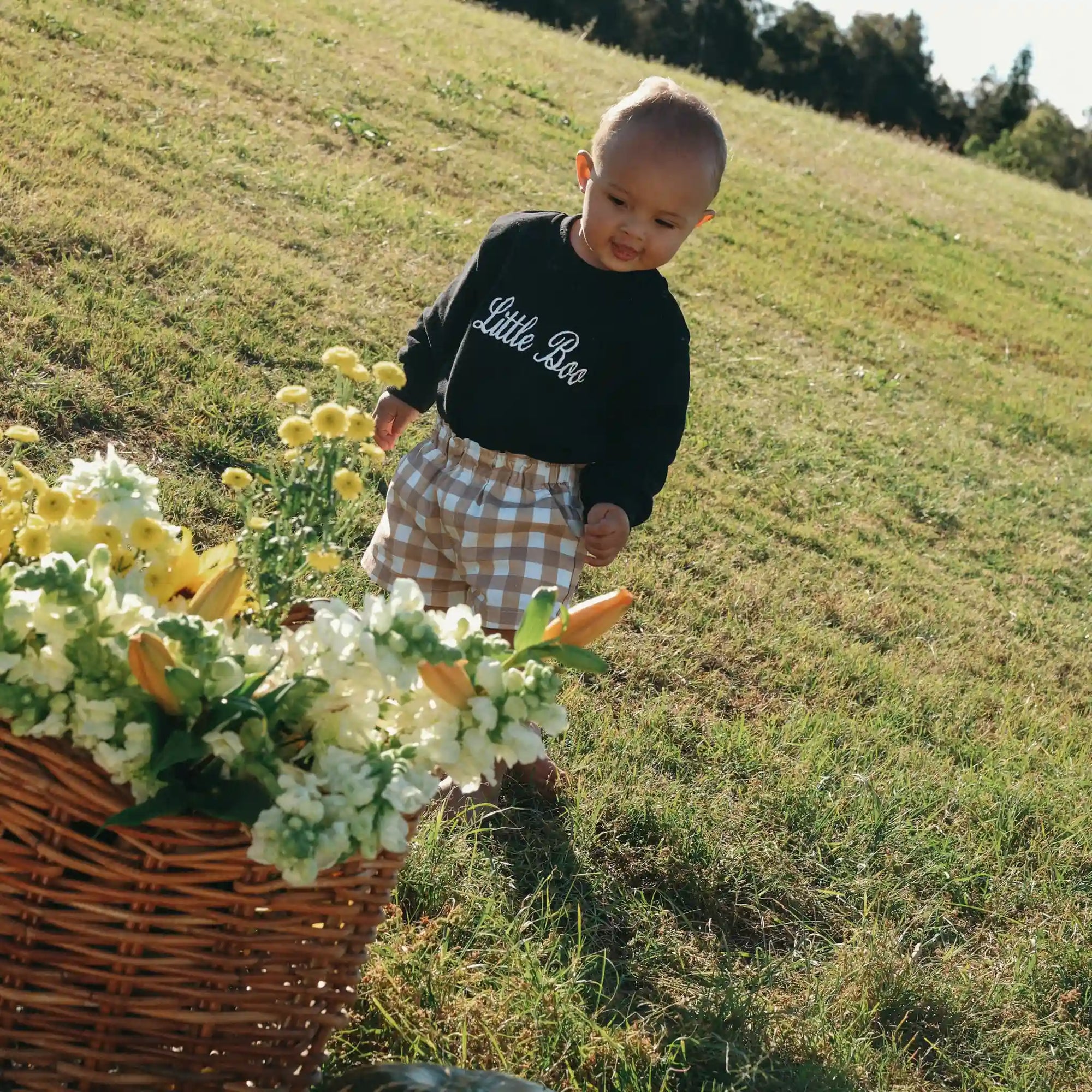 Baby in a black Little Boo sweatshirt and tan gingham shorts walking beside a basket of yellow flowers